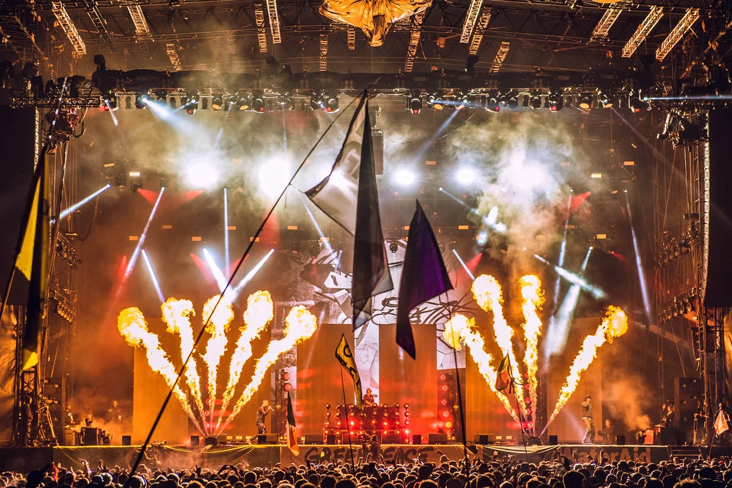 A lively concert stage with bright lights, pyrotechnics, and smoke effects, viewed from the audience, with flags waving in the foreground.