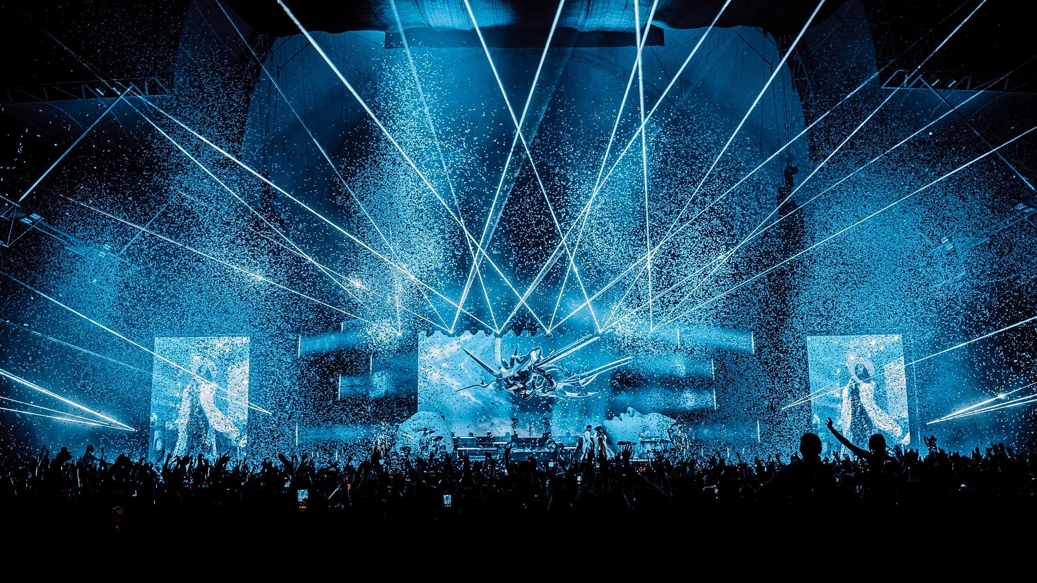 A concert scene with a stage illuminated by blue laser lights and confetti, crowd silhouettes dancing and raising hands.