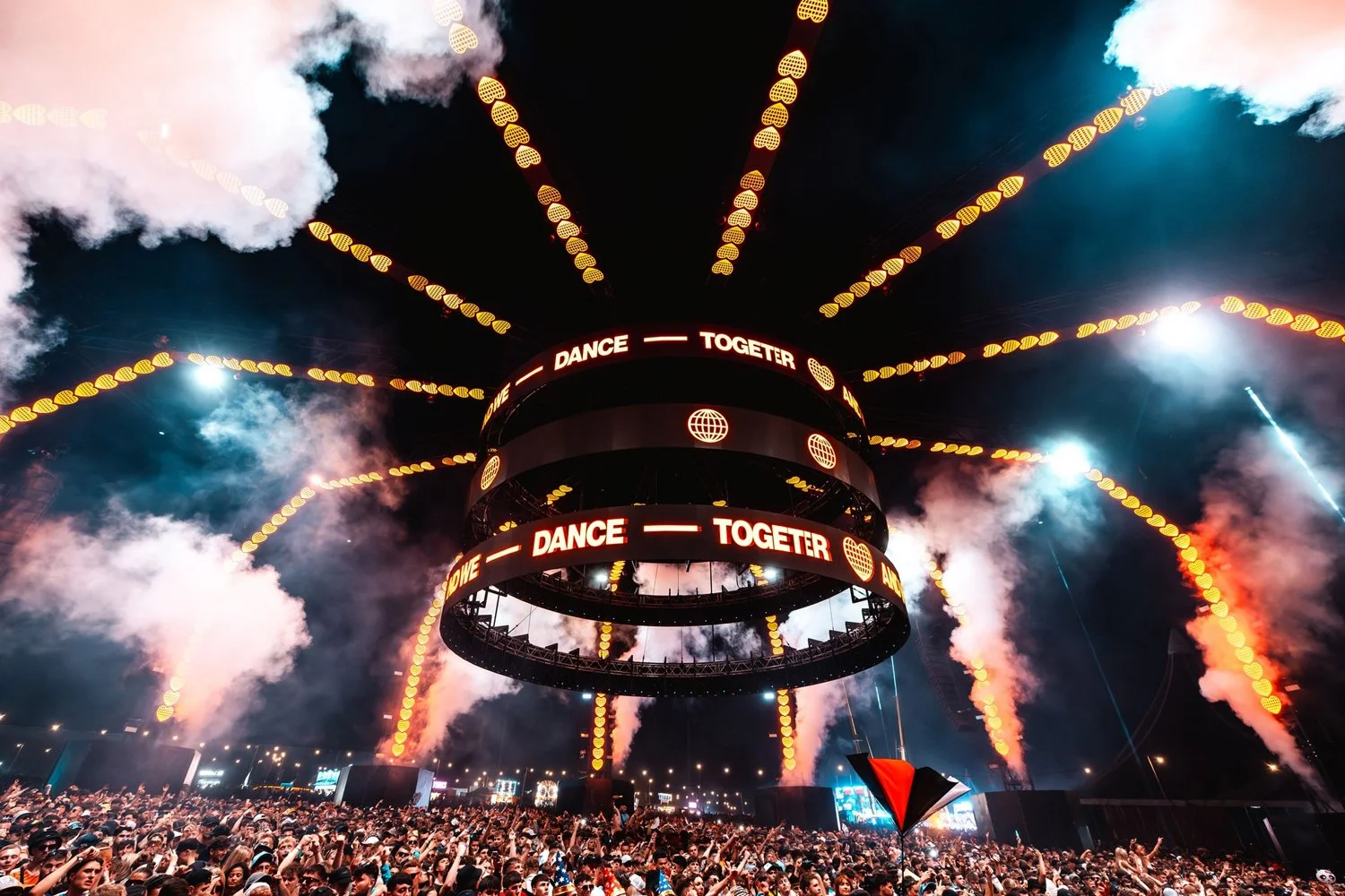 A large crowd at an outdoor music festival or concert with a stage and a large circular display. The display shows the words 'DANCE TOGETHER', surrounded by floating heart and globe icons, and there are bright lights and smoke effects.