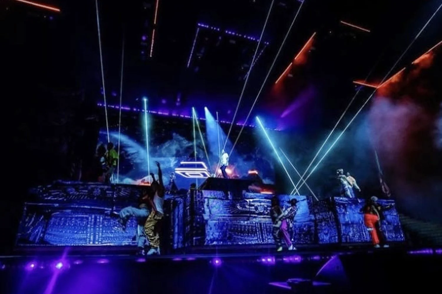 A live concert scene with performers on a futuristic stage illuminated by blue and purple lights, with laser beams projecting across the stage and fog effects adding to the vibrant atmosphere.