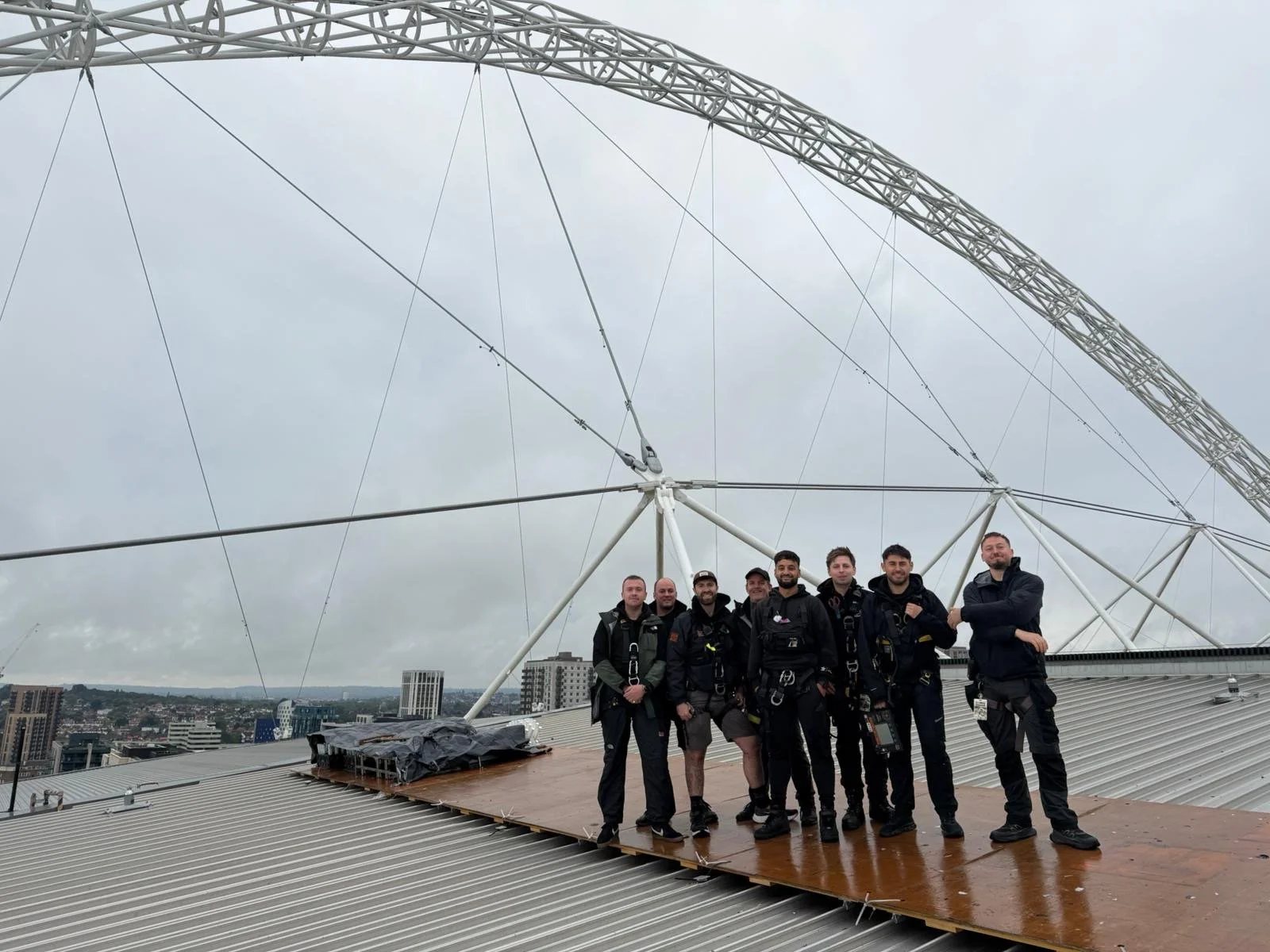 Eight people standing on a rooftop with a large Ferris wheel structure behind them, city skyline visible in the distance, cloudy sky overhead.
