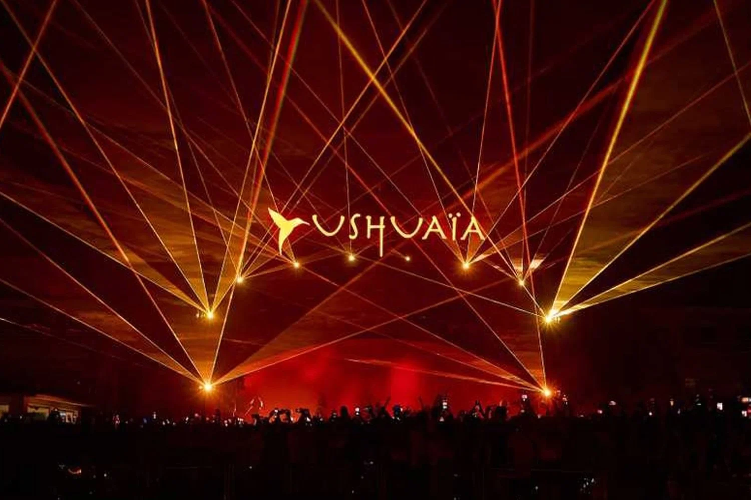A concert with bright yellow and orange laser lights in a dark venue. The sign 'USHUAIA' with a stylized yellow hummingbird is displayed in the center.
