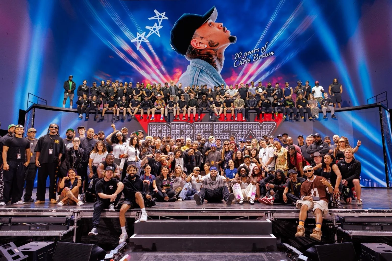 Group photo of people on stage during a celebration or concert, with a large screen in the background displaying a portrait of a man and the text '20 Years of Chris Brown'.