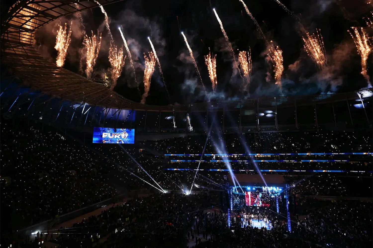 A nighttime view of a large indoor sports arena with fireworks in the sky, a boxing ring in the center, and spectators watching the event.