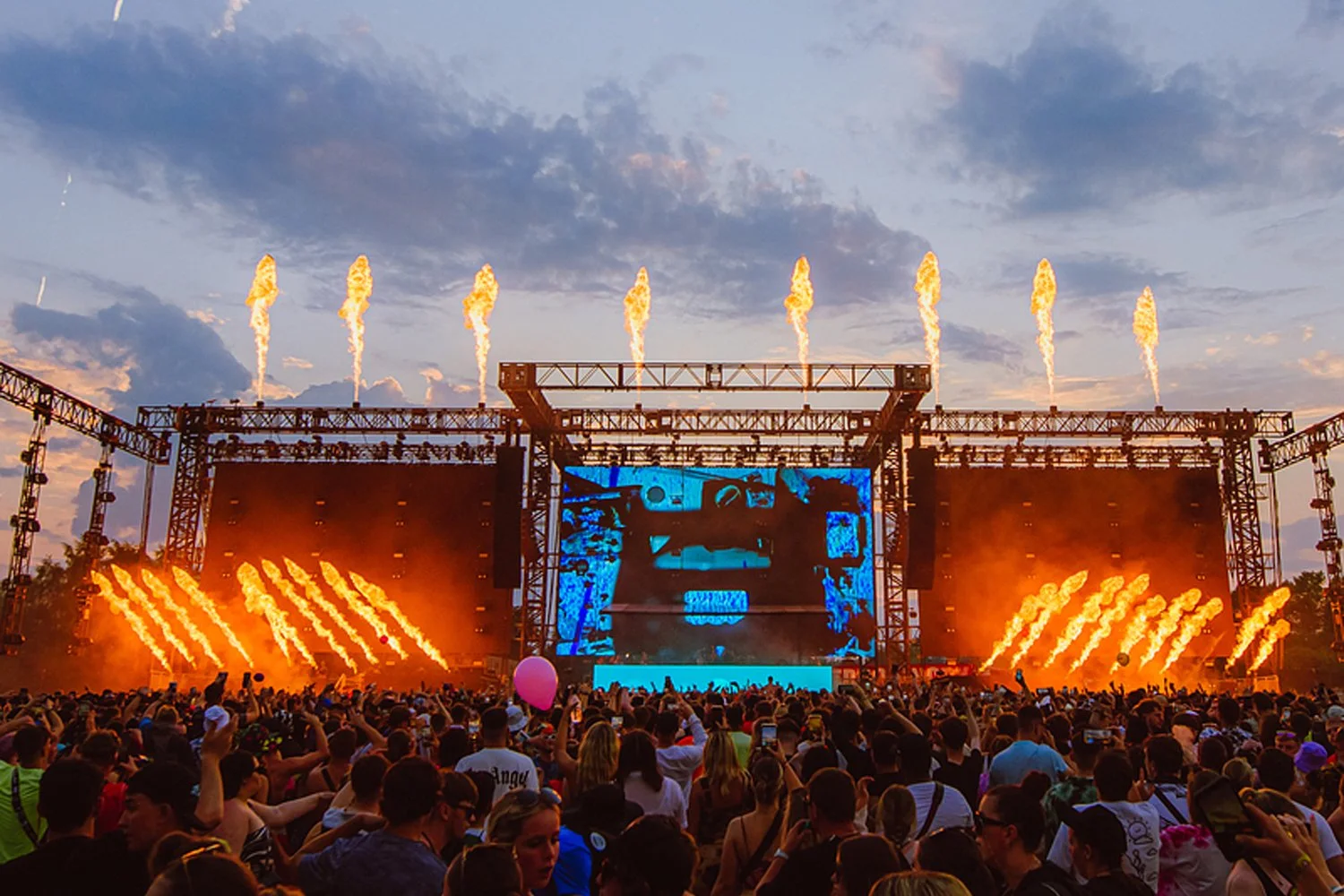 A large outdoor music festival stage with fireworks and pyrotechnics, surrounded by a crowd of festival-goers during sunset.
