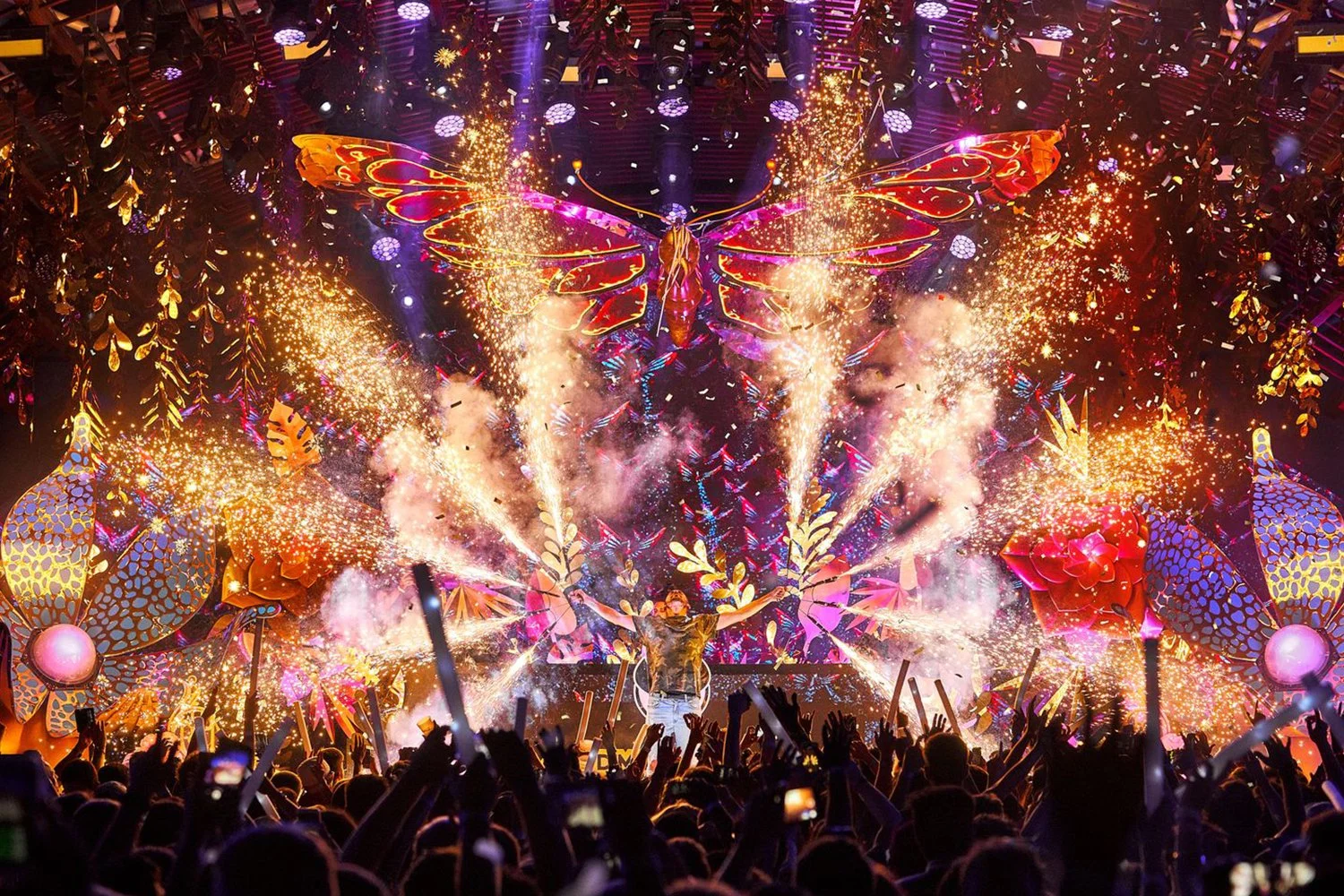 A concert stage illuminated with vibrant lights, fireworks, and sparkling effects, featuring a performer with arms raised in front of a large, colorful butterfly and floral decorations, surrounded by an audience.
