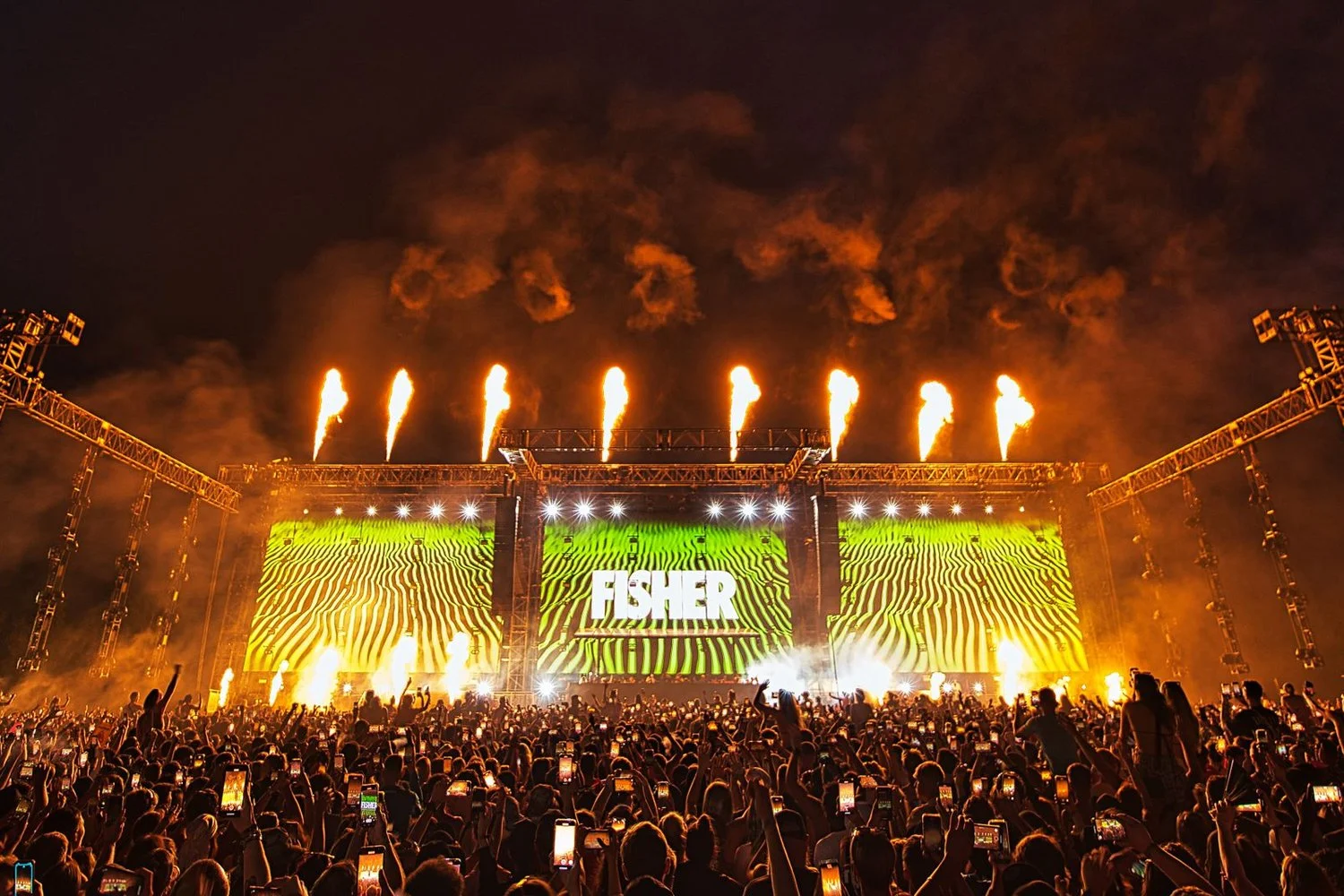 A large outdoor concert at night with a stage illuminated in green and yellow, the word 'FISHER' displayed on a screen, fireworks and flames shooting into the sky, and a crowd of fans taking photos and videos.