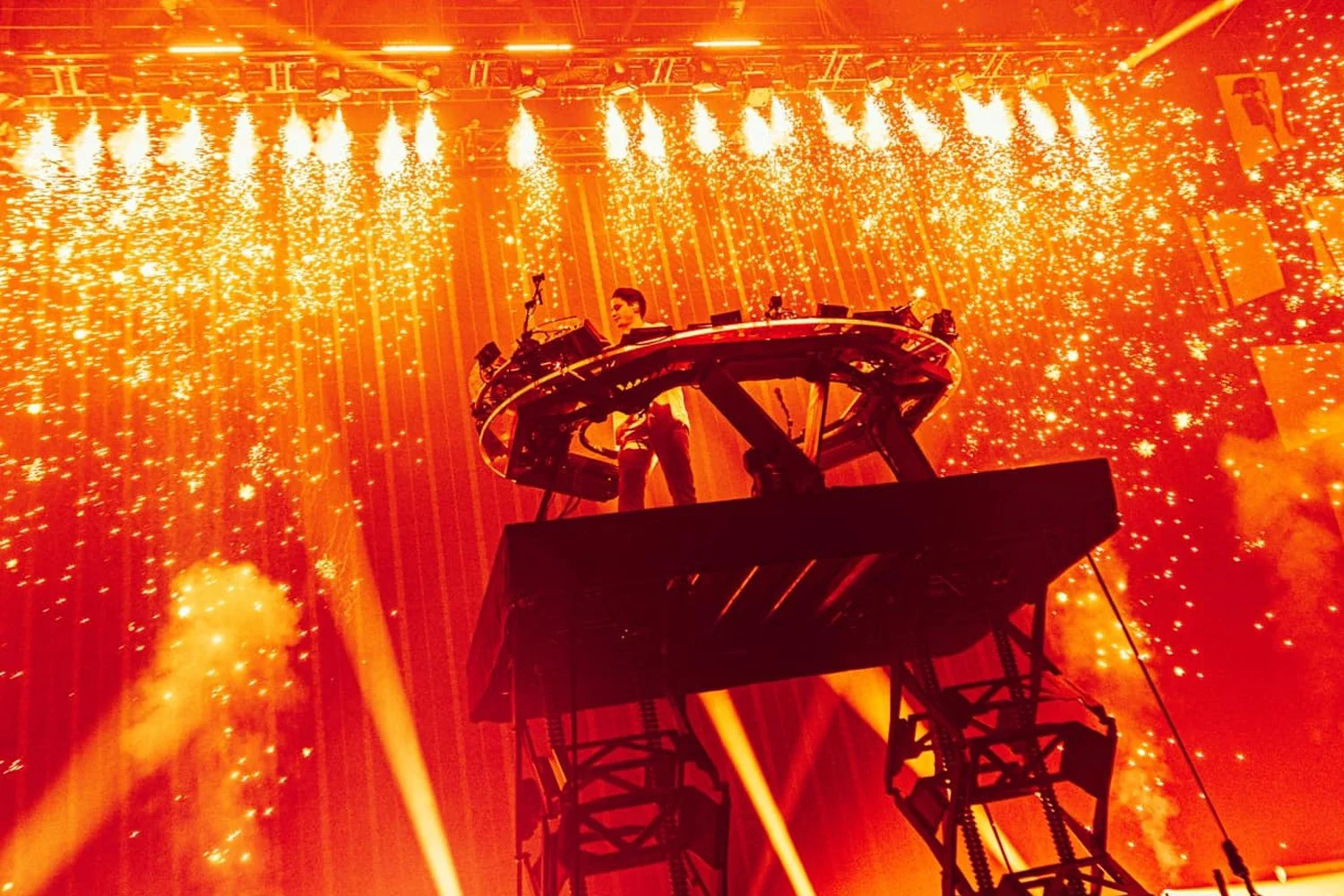 A DJ performs on a circular platform elevated above the stage, surrounded by fireworks and bright orange lighting at a concert or festival.