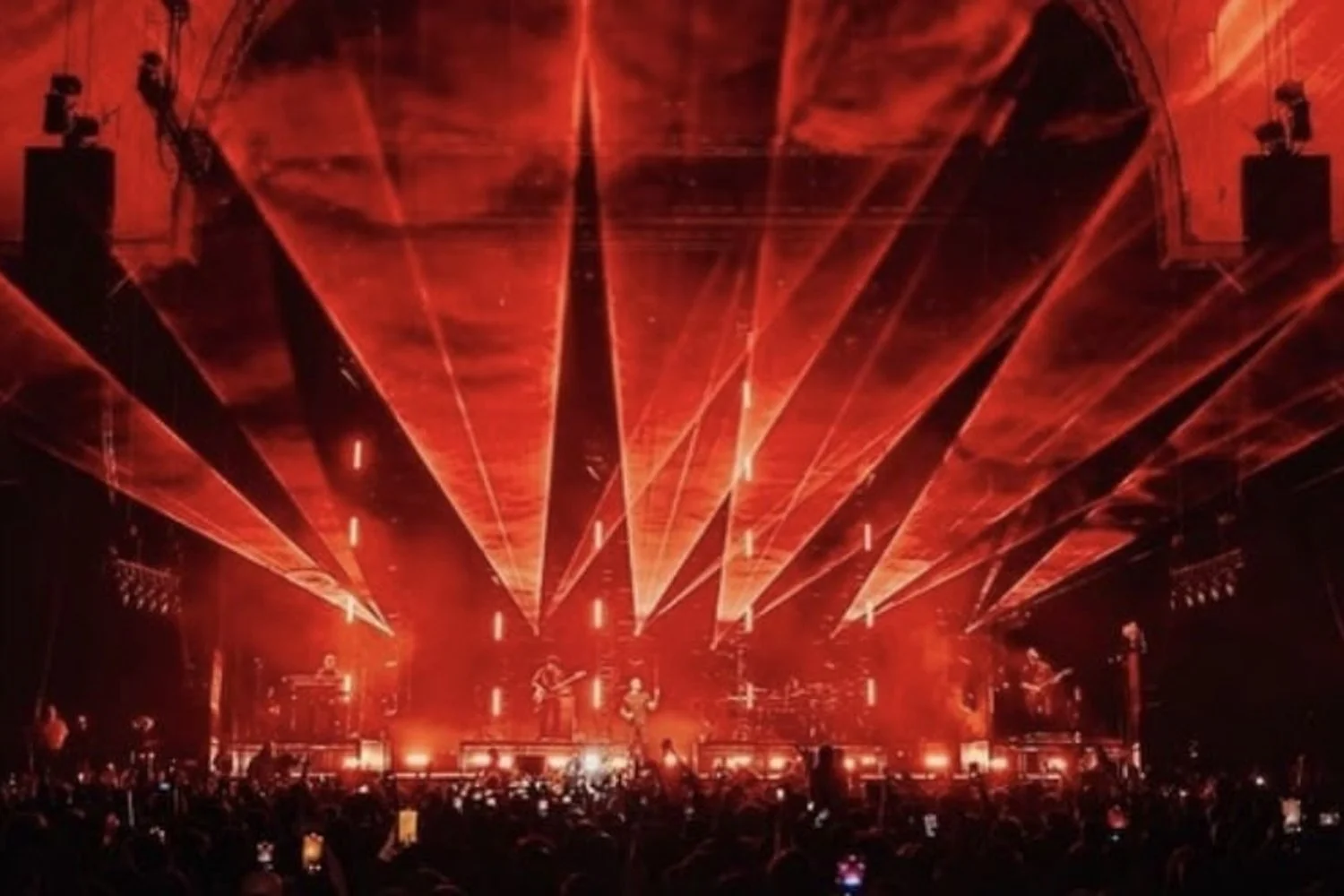 Concert stage with red laser light show, musicians playing on stage, surrounded by an audience taking photos.