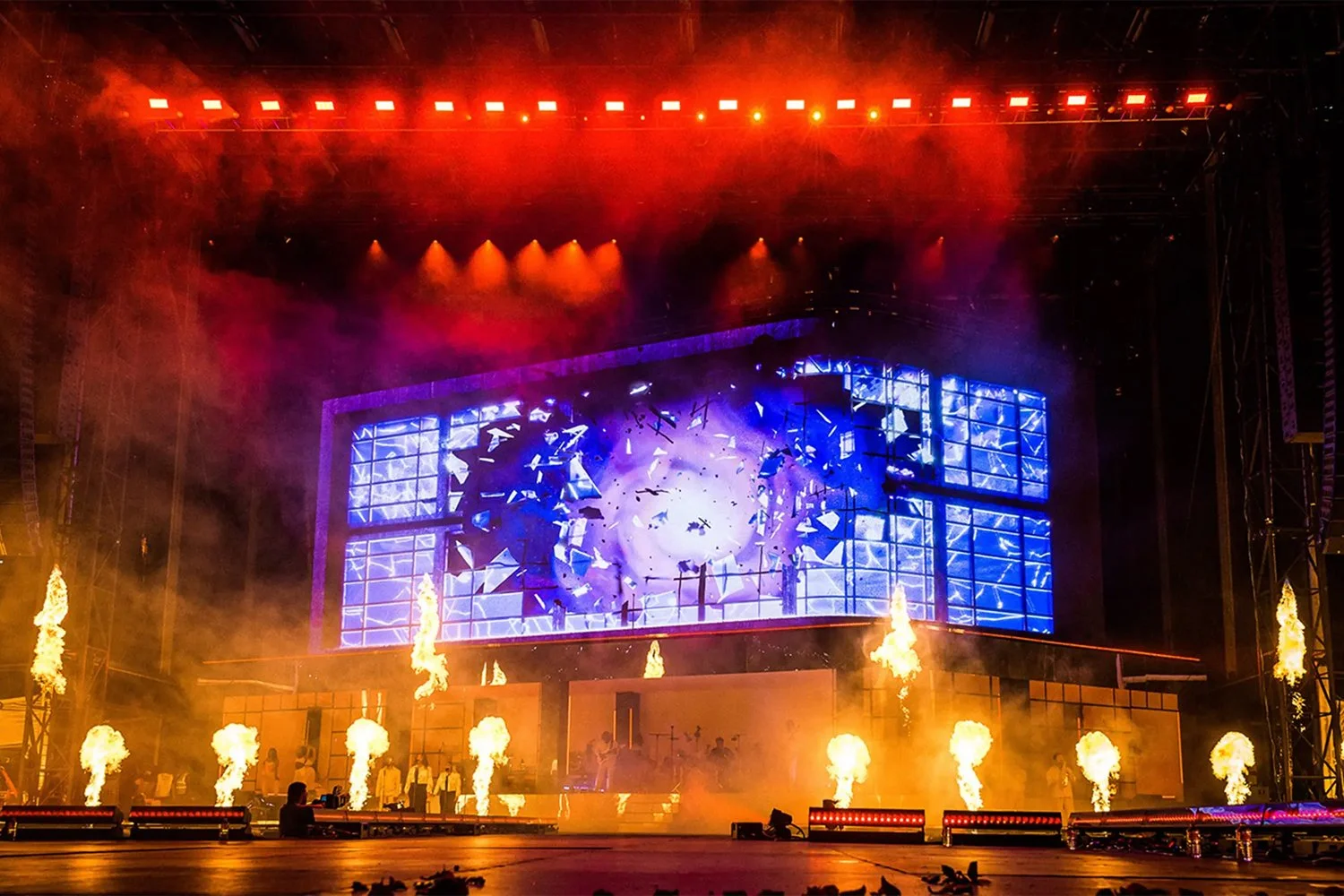 A concert stage with bright lights, pyrotechnics, and a large digital screen displaying abstract visuals, with performers and equipment visible in the background.