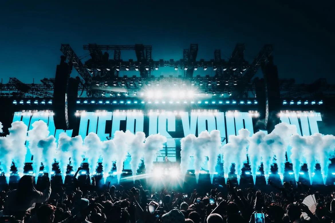 Music concert with bright stage lights, fog effects, and a crowd of people taking photos.