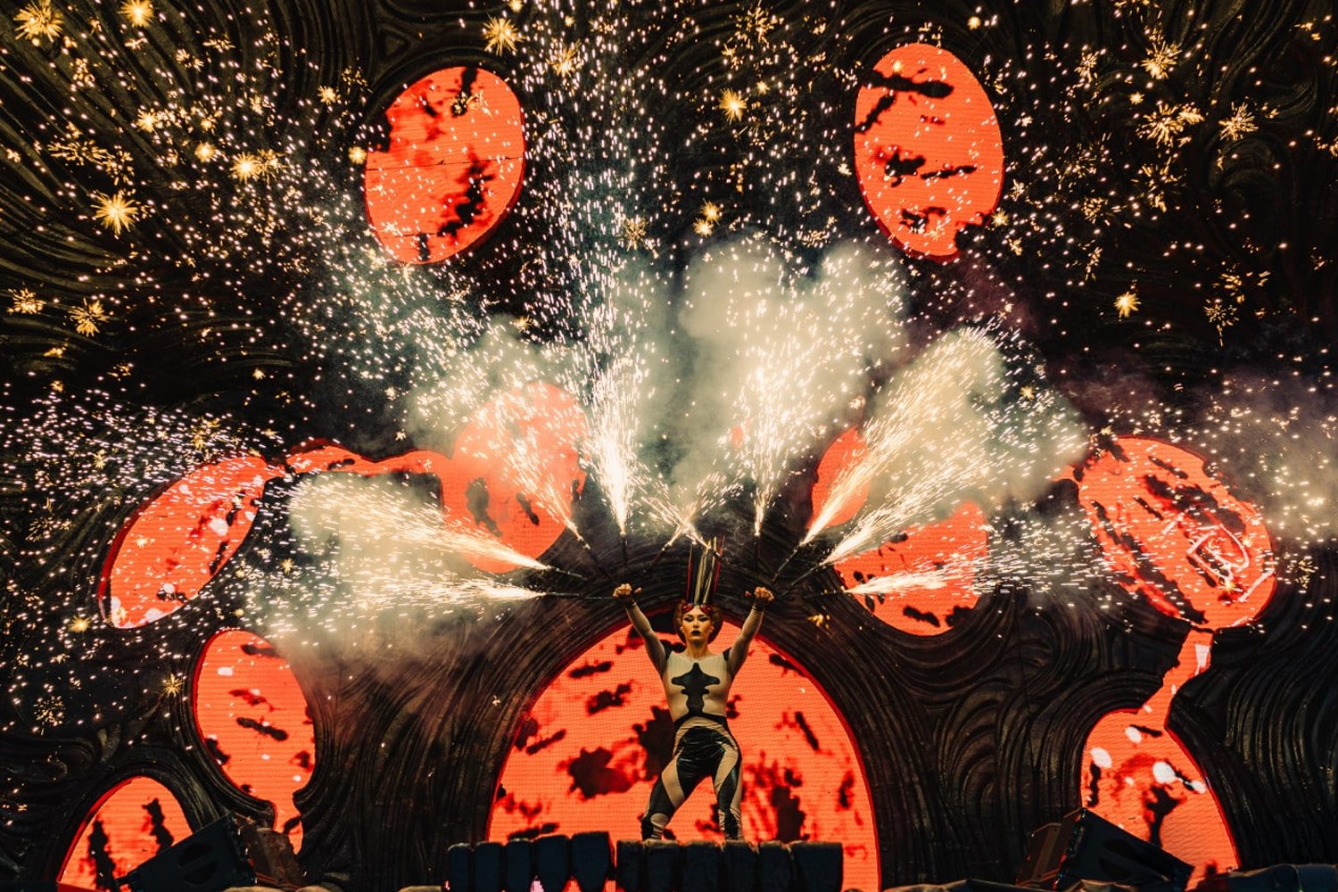 A performer on stage with arms raised, holding sparklers, with a backdrop of large digital screens displaying red, bat-shaped images, and fireworks and sparks overhead.