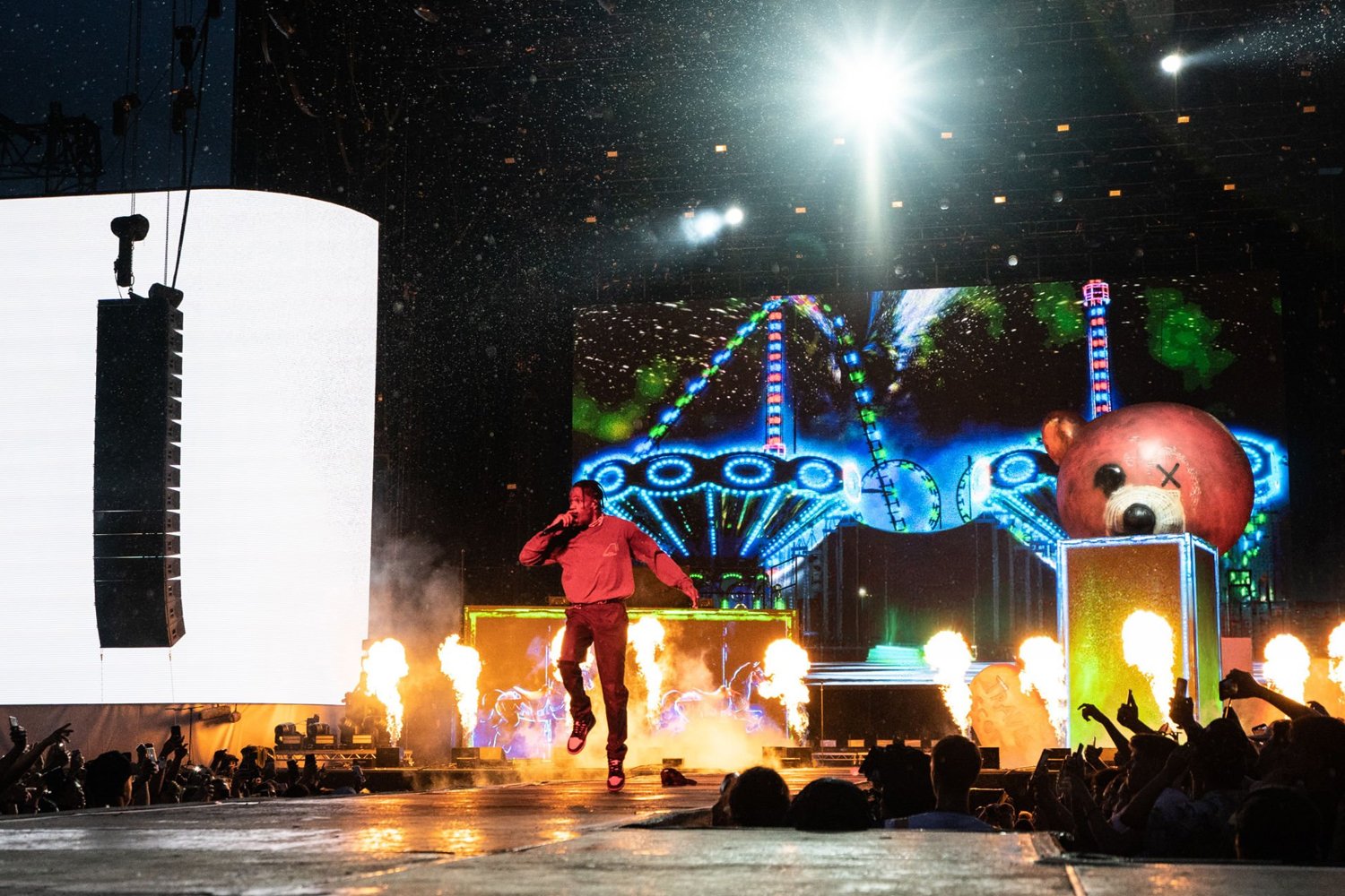 Performer on stage at a concert, with an amusement park and a large bear sculpture in the background, flames and bright lights creating a lively atmosphere.