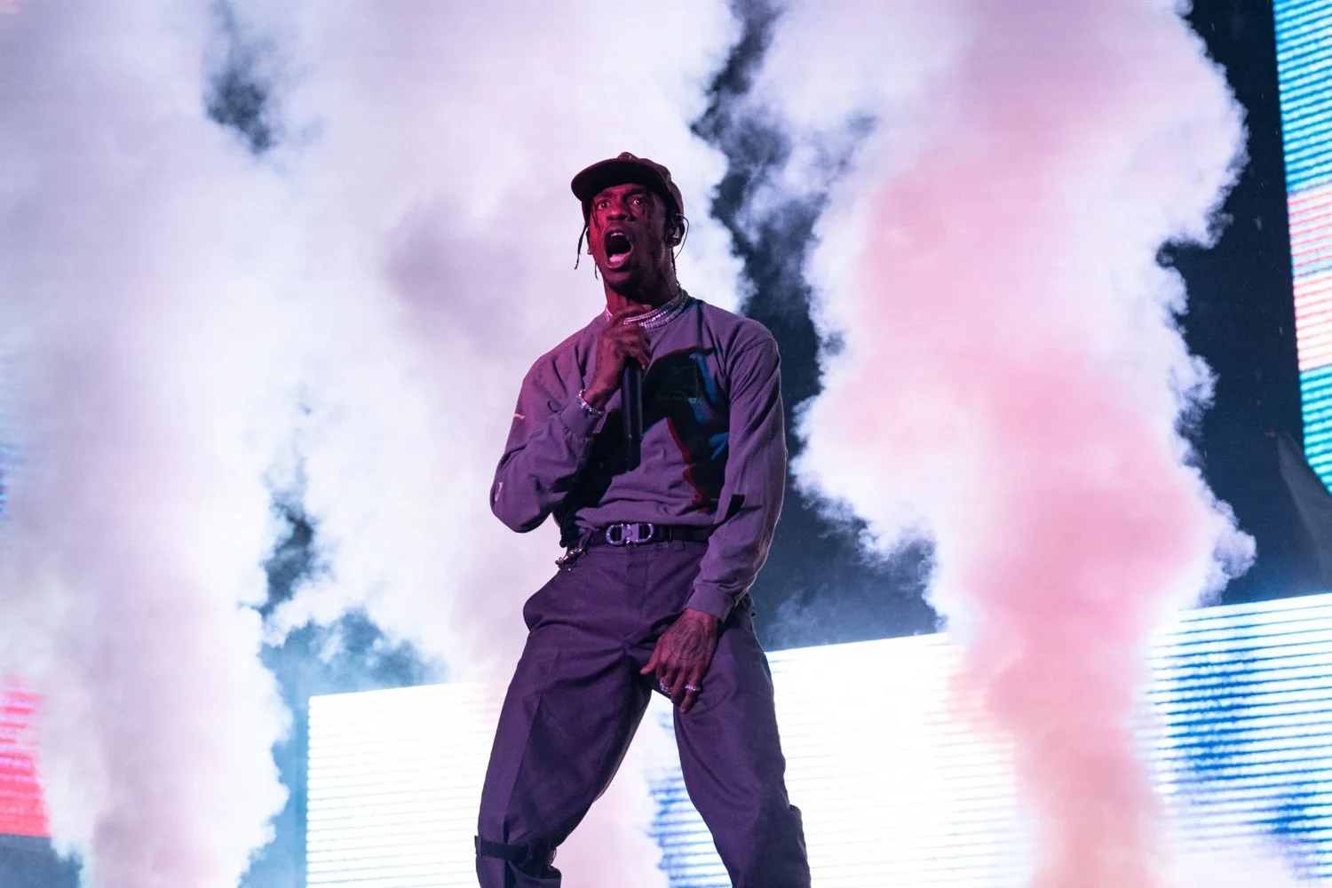 A male performer on stage with smoke and colorful lights in the background, holding a microphone, with an expressive facial expression.