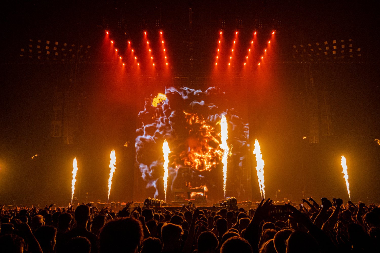 A concert stage with pyrotechnics and fire effects, crowd in the foreground, bright red and orange lights, smoke, and flames.