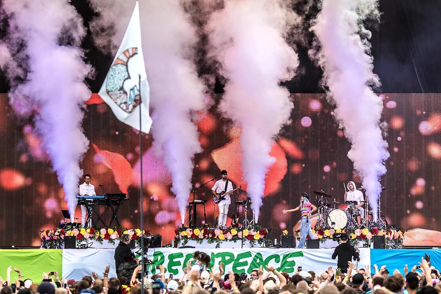 A band performing on stage with colorful smoke effects, a large digital background, and a crowd in front. The stage is decorated with flowers and has a banner reading 'PEACE' in large green letters.