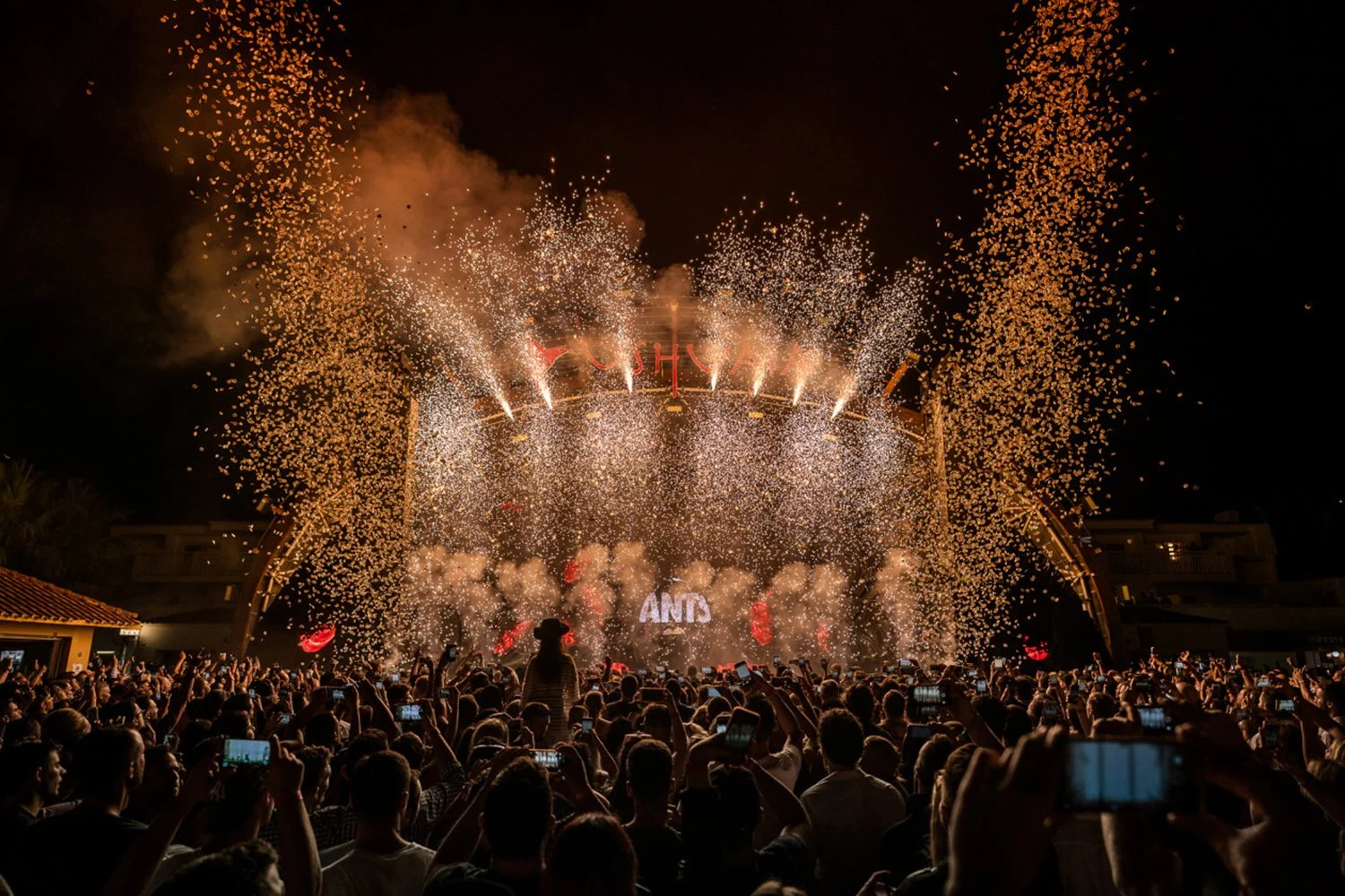 Crowd watching fireworks and confetti at a nighttime concert or festival, stage with the word 'ANIS', fireworks in the sky, many audience members taking photos with smartphones.