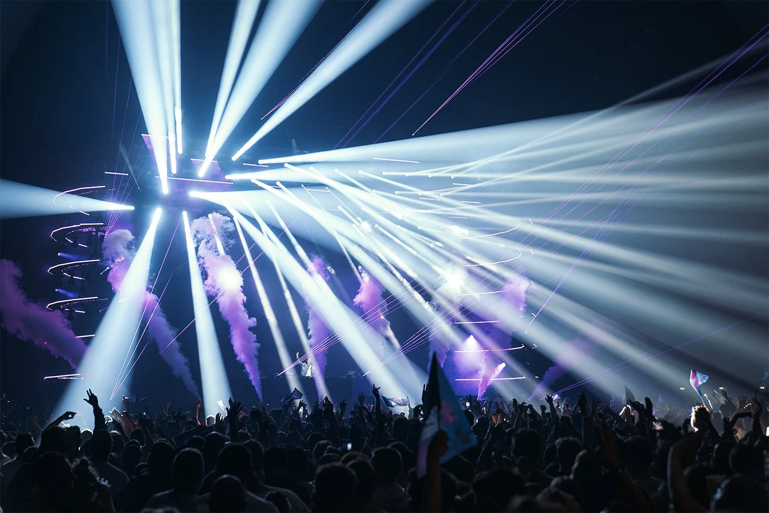 A concert crowd with hands raised, bright white and purple laser lights and smoke effects in a dark indoor venue.