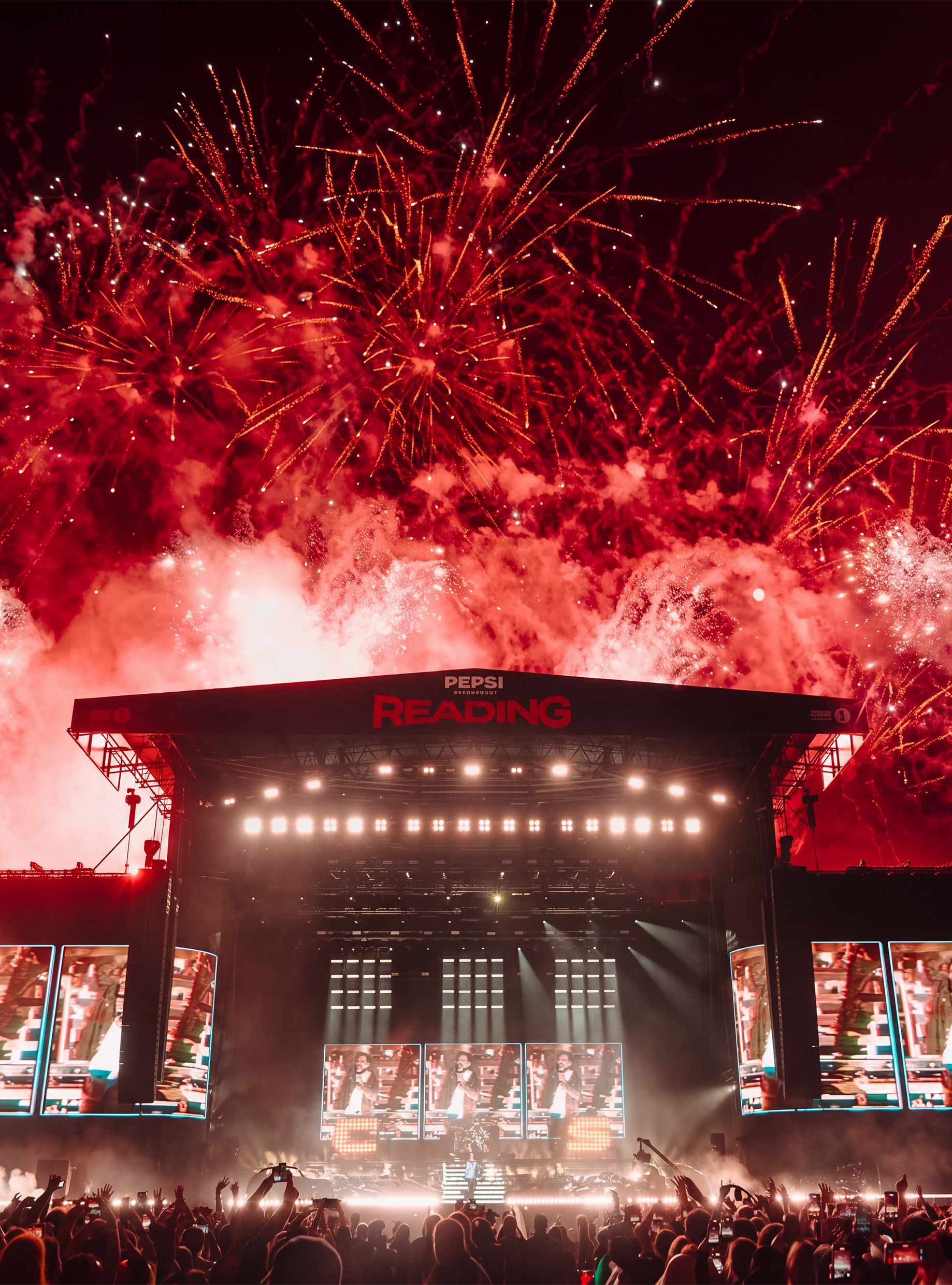 Fireworks explode in the night sky above a large outdoor concert stadium filled with a crowd, with a stage featuring large screens and bright lights.