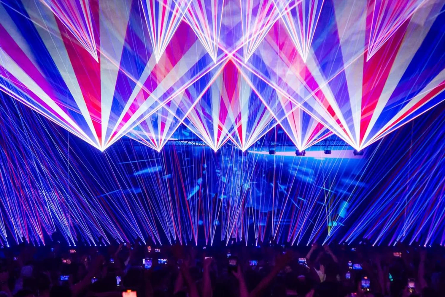 Colorful laser light show with beams of purple, pink, and blue lights crisscrossing above an audience at a concert or event.