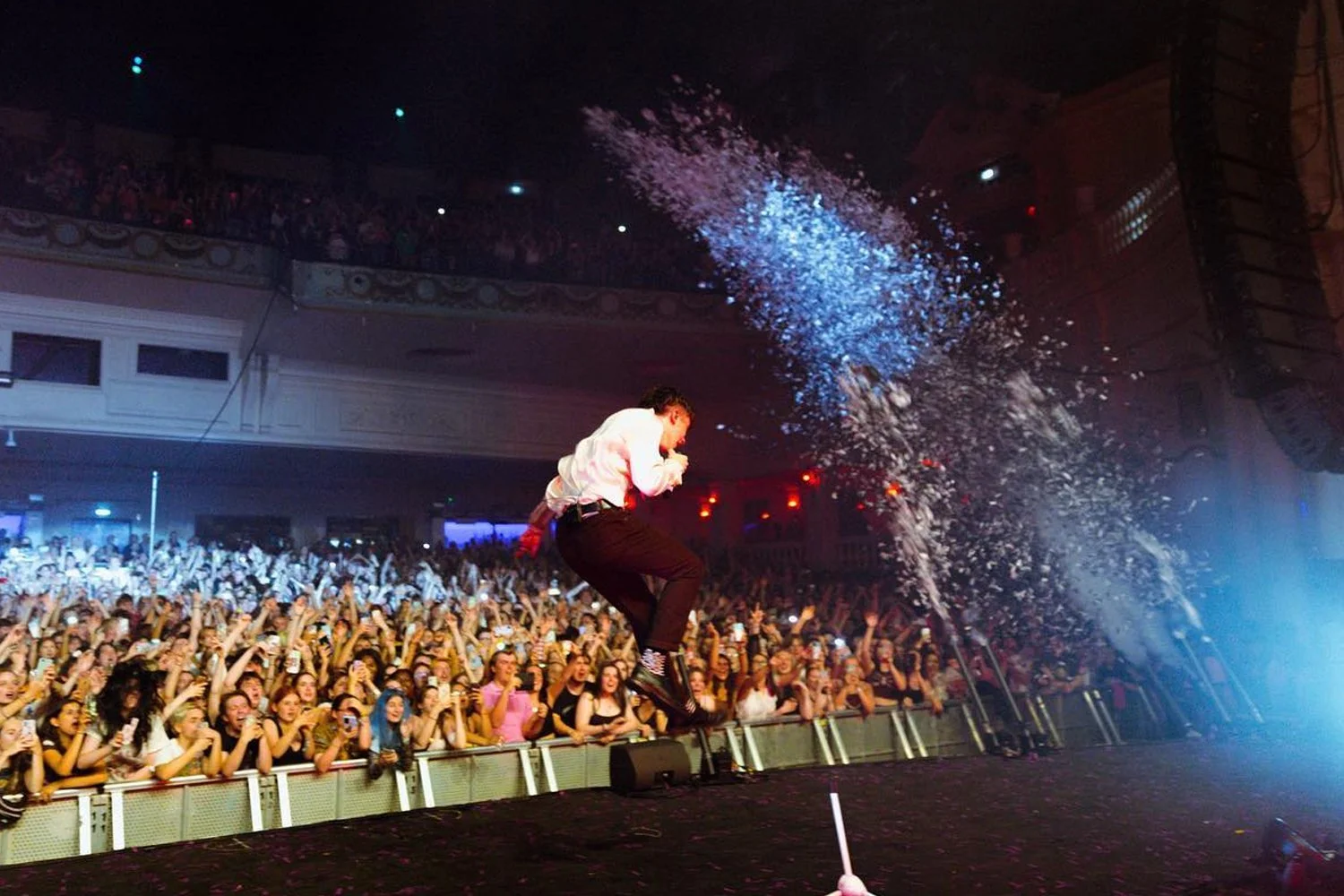 A performer in a white shirt and black pants is jumping and throwing confetti into the air during a concert, with a cheering crowd in the background.