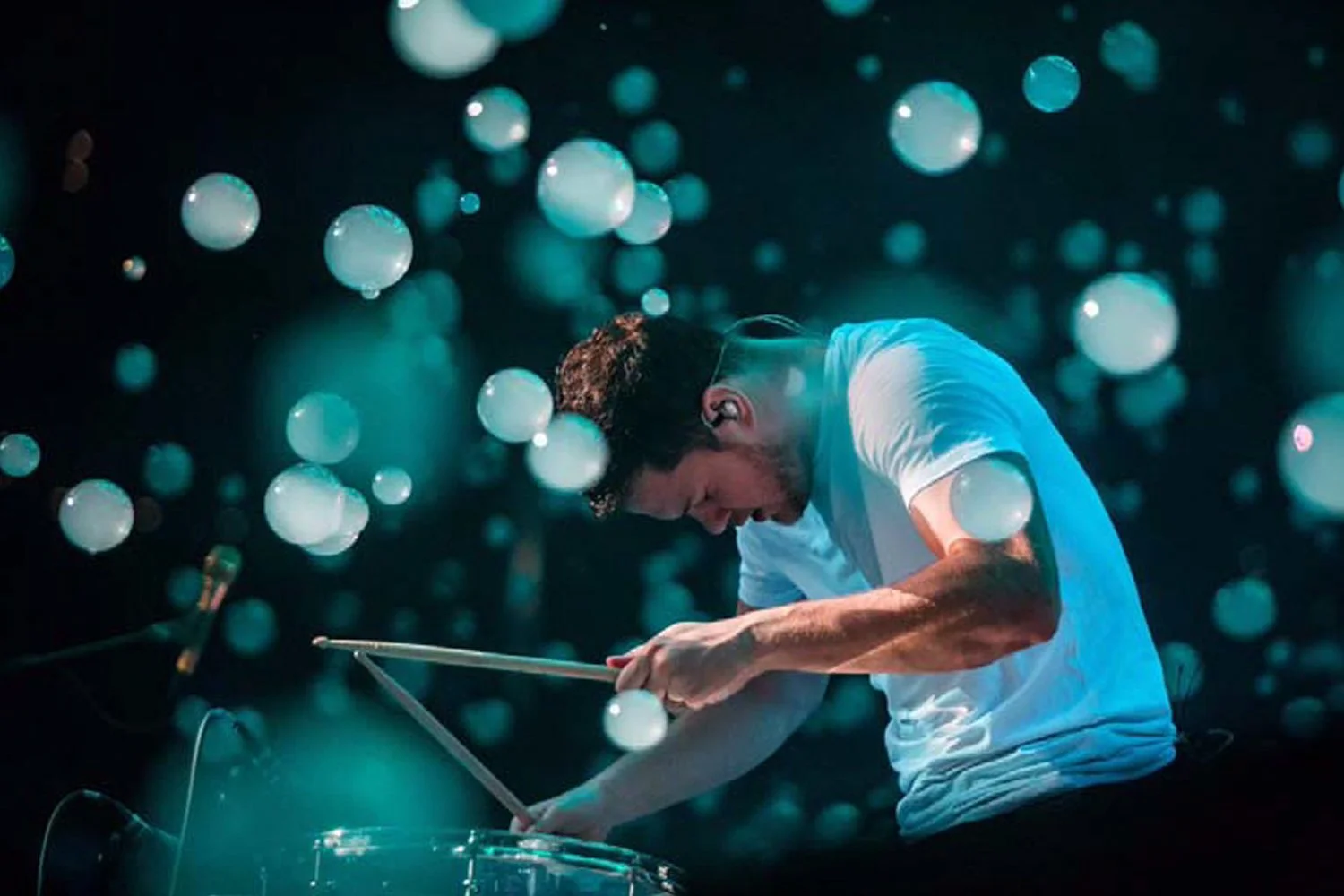 A young man in a white t-shirt is playing drums passionately, surrounded by floating glowing orbs of light in a dark setting.