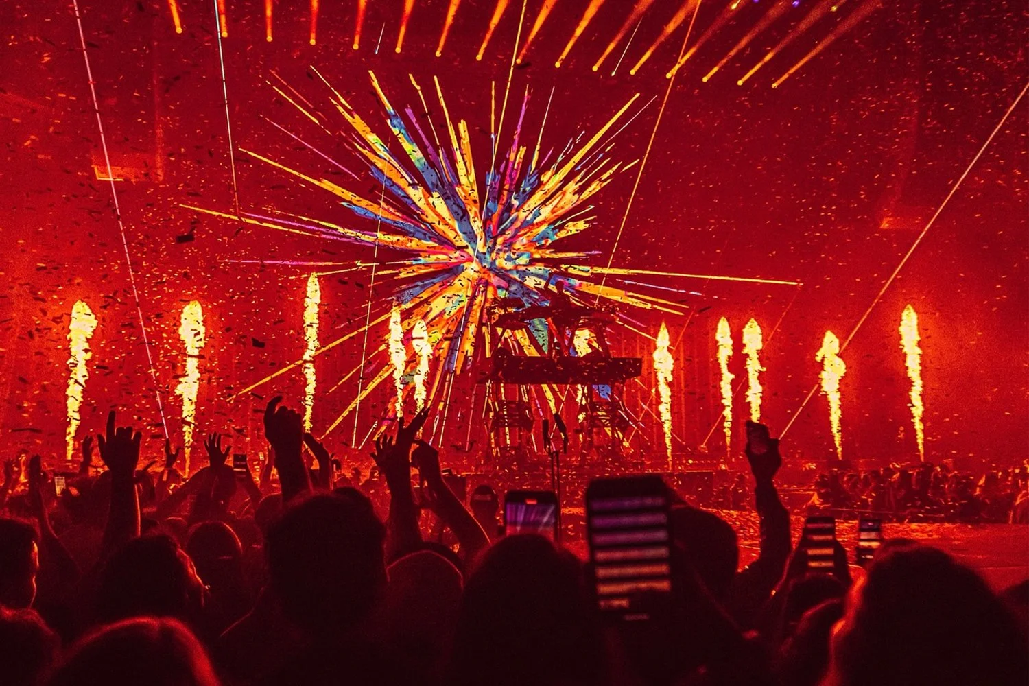 Crowd at a concert or festival with fireworks and bright lights illuminating the night sky, some people holding up phones to record the event.