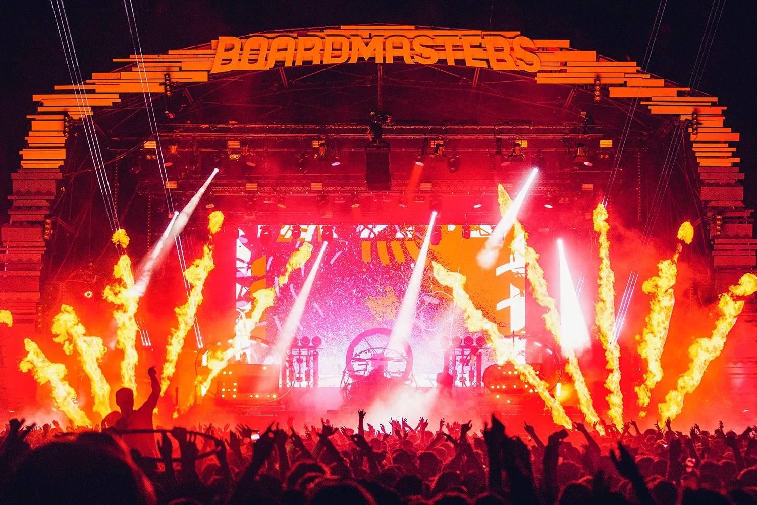 A concert at Boardmasters festival with a stage, red lighting, flames, and a crowd of people dancing and raising their hands.