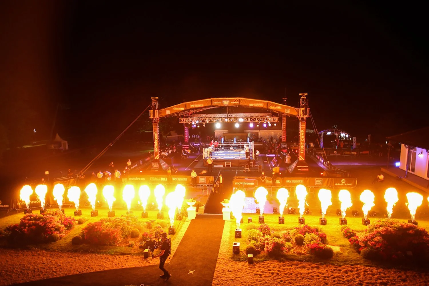 Nighttime boxing match setup with a boxing ring on a stage and pyrotechnics in front of the stage, surrounded by flowers and lights.