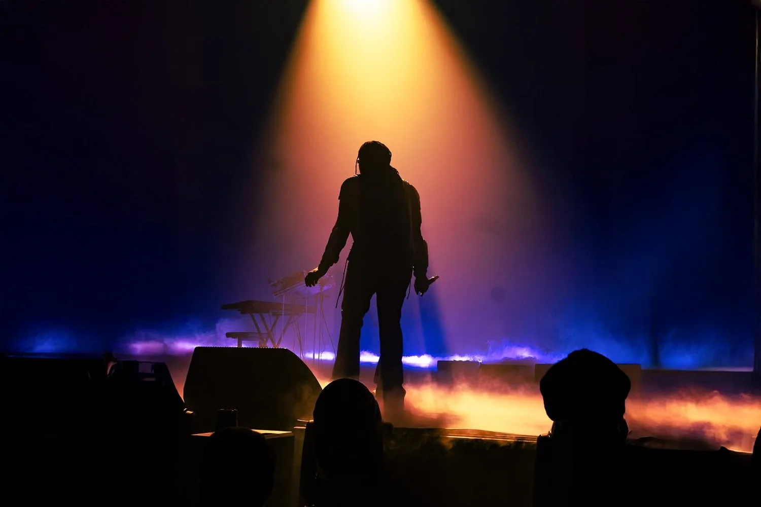 Silhouette of a person standing on a stage with a keyboard, holding a microphone, illuminated by colorful stage lights with purple and blue hues, and a yellow spotlight overhead.