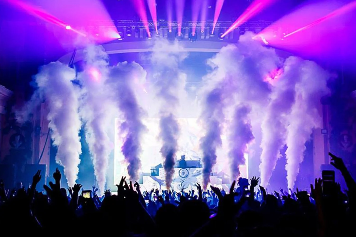 A concert scene with bright purple and white stage lights, smoke effects, and a crowd of people with hands raised.