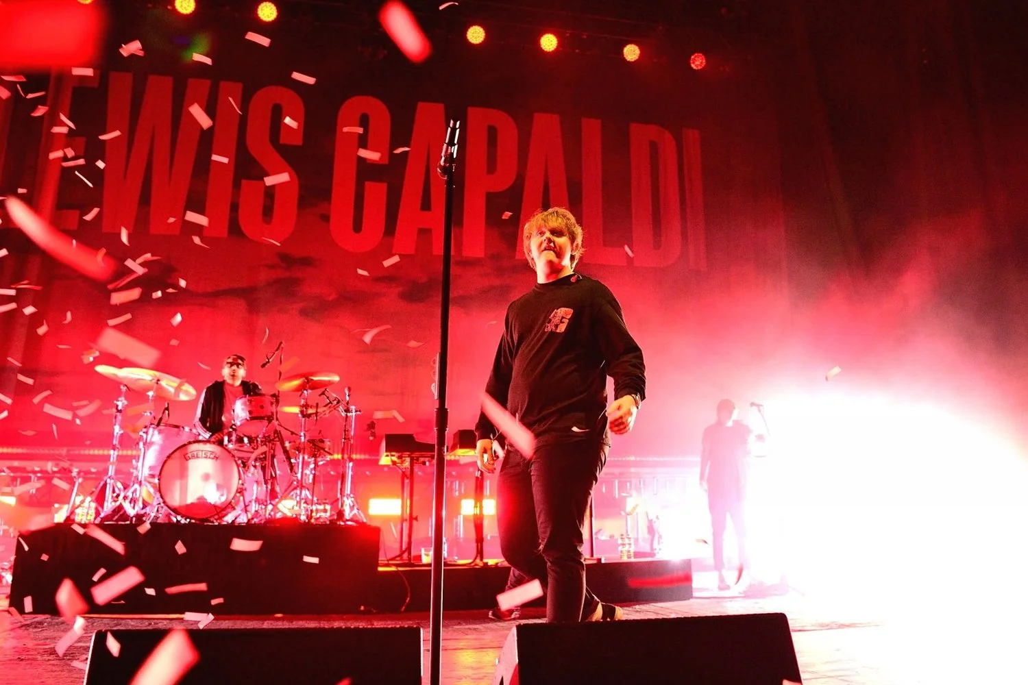 A live music performance on stage with a band, with red lighting and confetti falling, and a large backdrop reading "WISH CAPITAL."