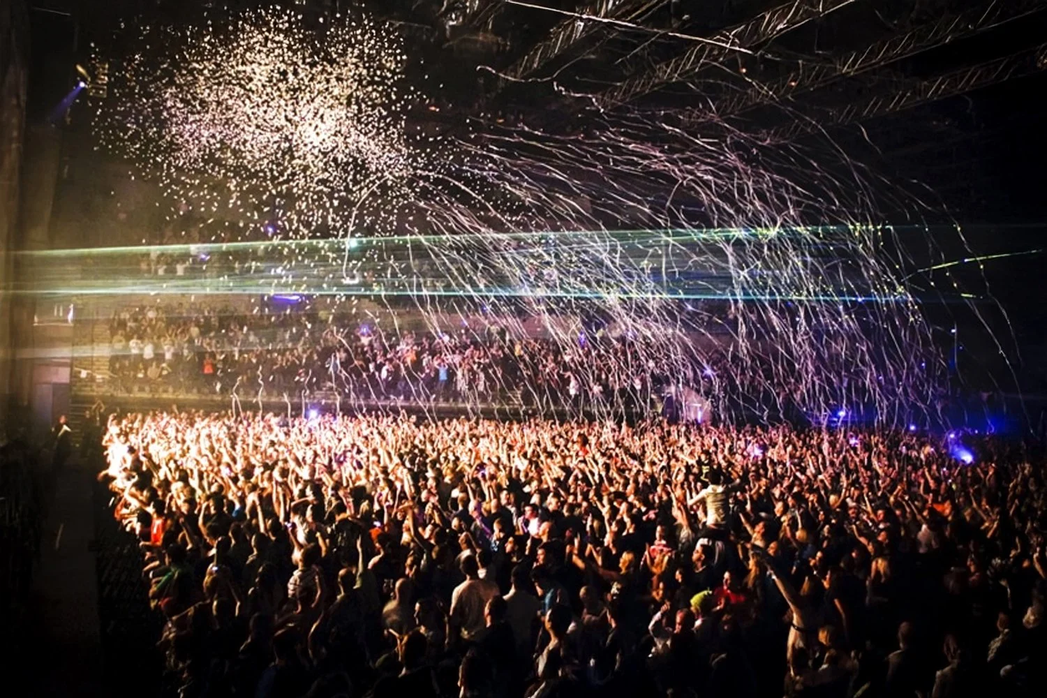 A large crowd of people at a concert or party with confetti and streamers flying through the air, illuminated by stage lights and fireworks.