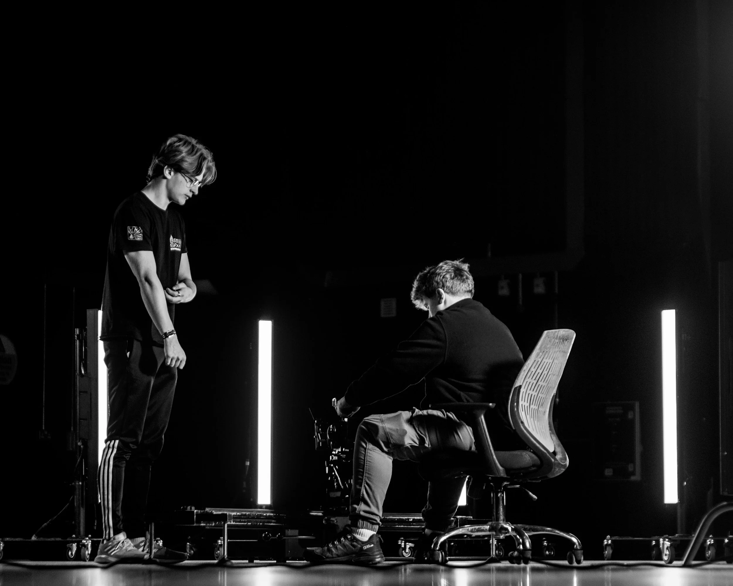 Two young men appear to be in a discussion or working together, with one standing and one sitting in a modern, dark room, illuminated by vertical light fixtures.