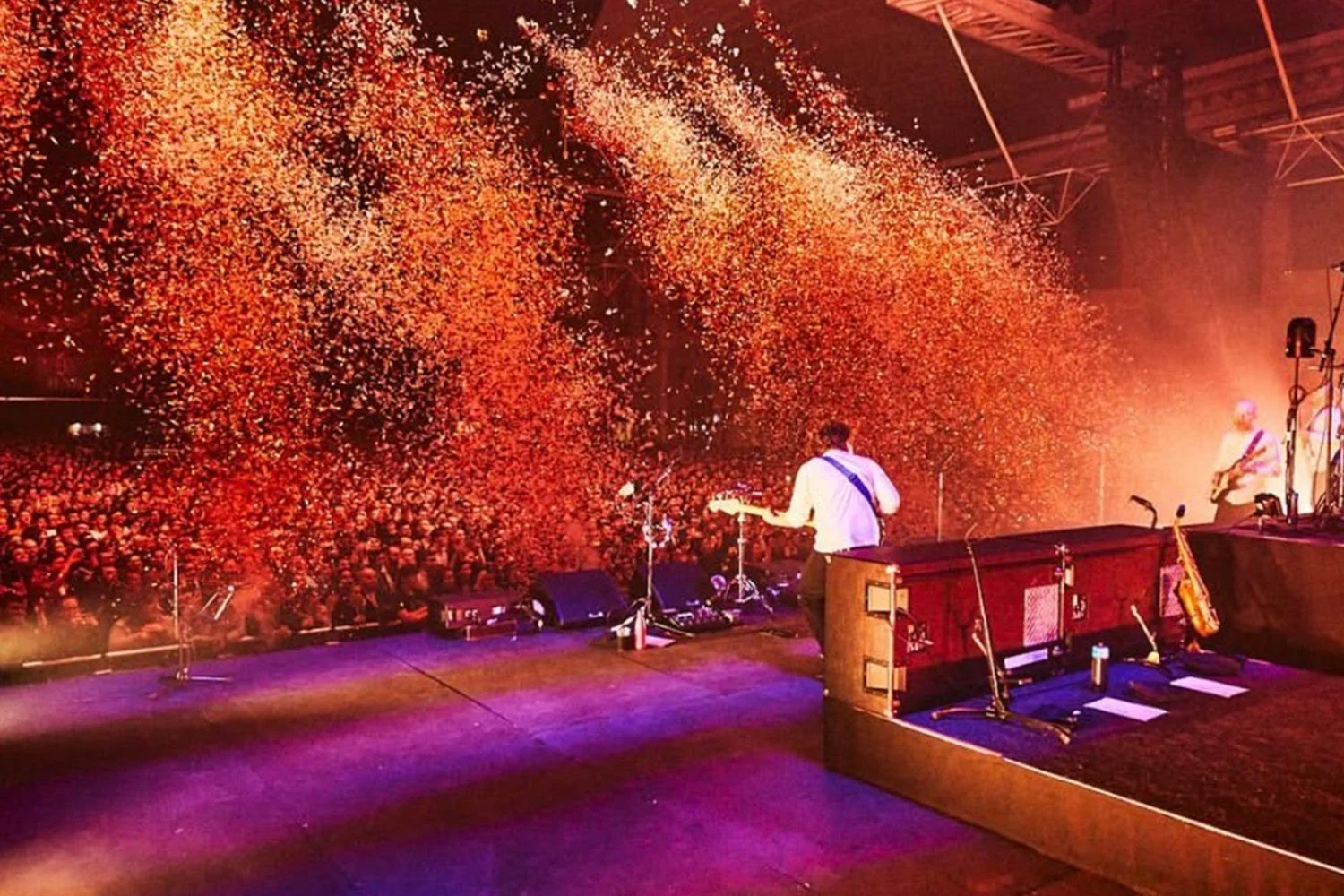 Live concert on stage with musicians playing guitars, with a large audience in front. Colorful confetti is falling from above as part of the celebration.