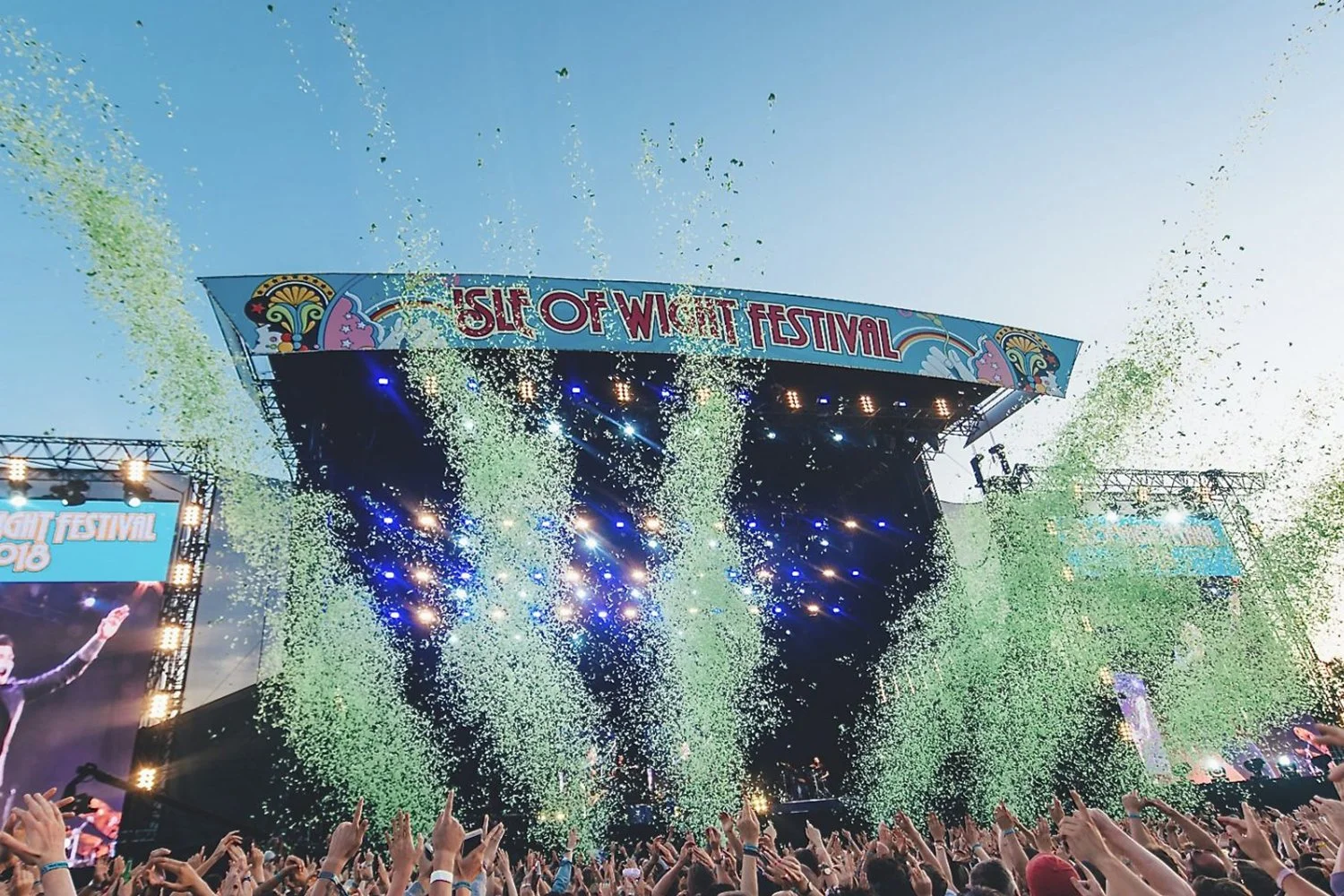 Crowd at a music festival with a large stage, colorful lights, and green confetti falling from above during a live performance at the Isle of Wight Festival.