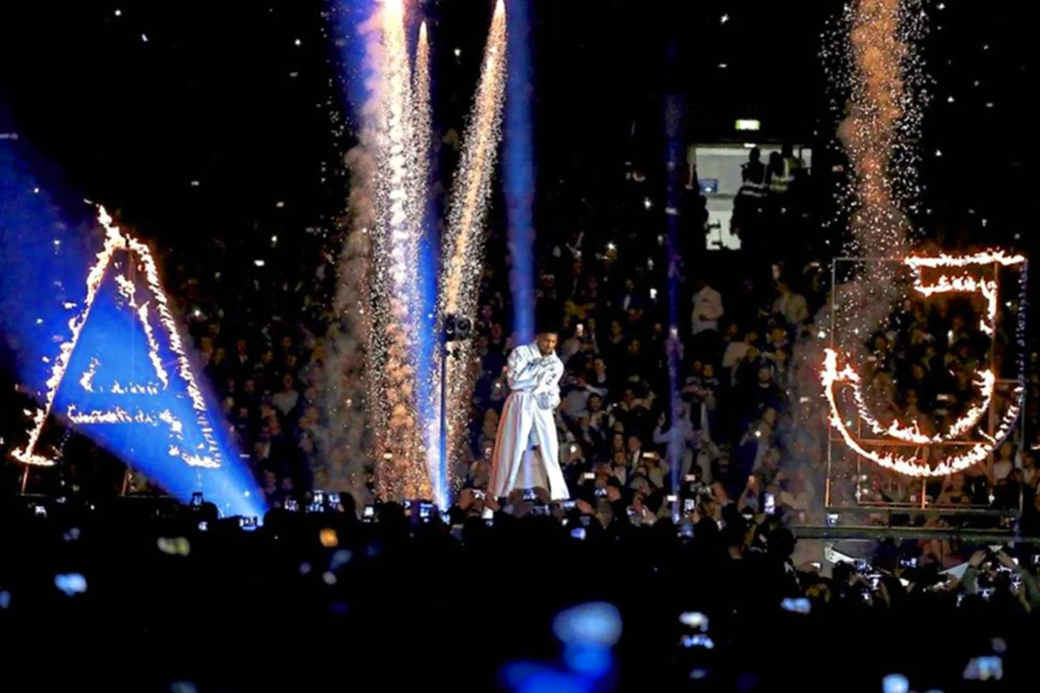 A large crowd watching a performance with fireworks and pyrotechnics, including a figure standing on stage surrounded by sparks and lasers, with symbols and flags illuminated in the background.