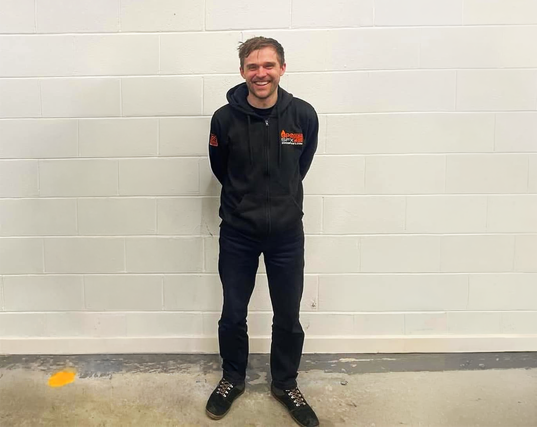A man standing and smiling in front of a white brick wall, wearing a black hoodie, dark pants, and sneakers.