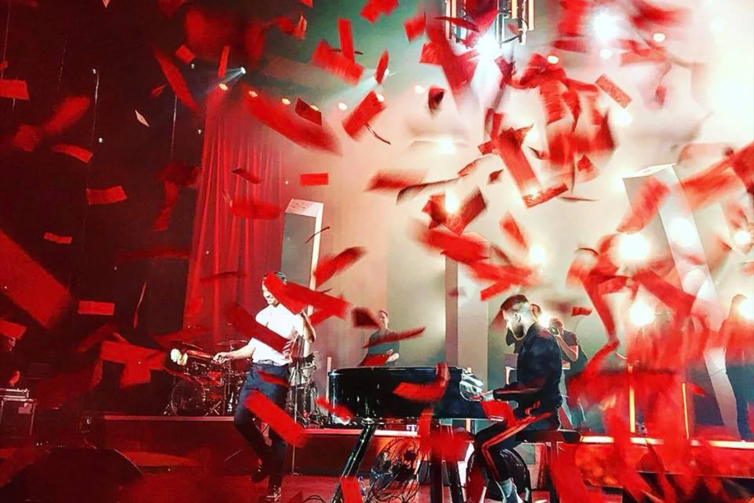 Musicians performing on stage with red confetti falling around them, including a pianist, drummer, and a person playing the keyboard, with a red curtain backdrop and stage lighting.