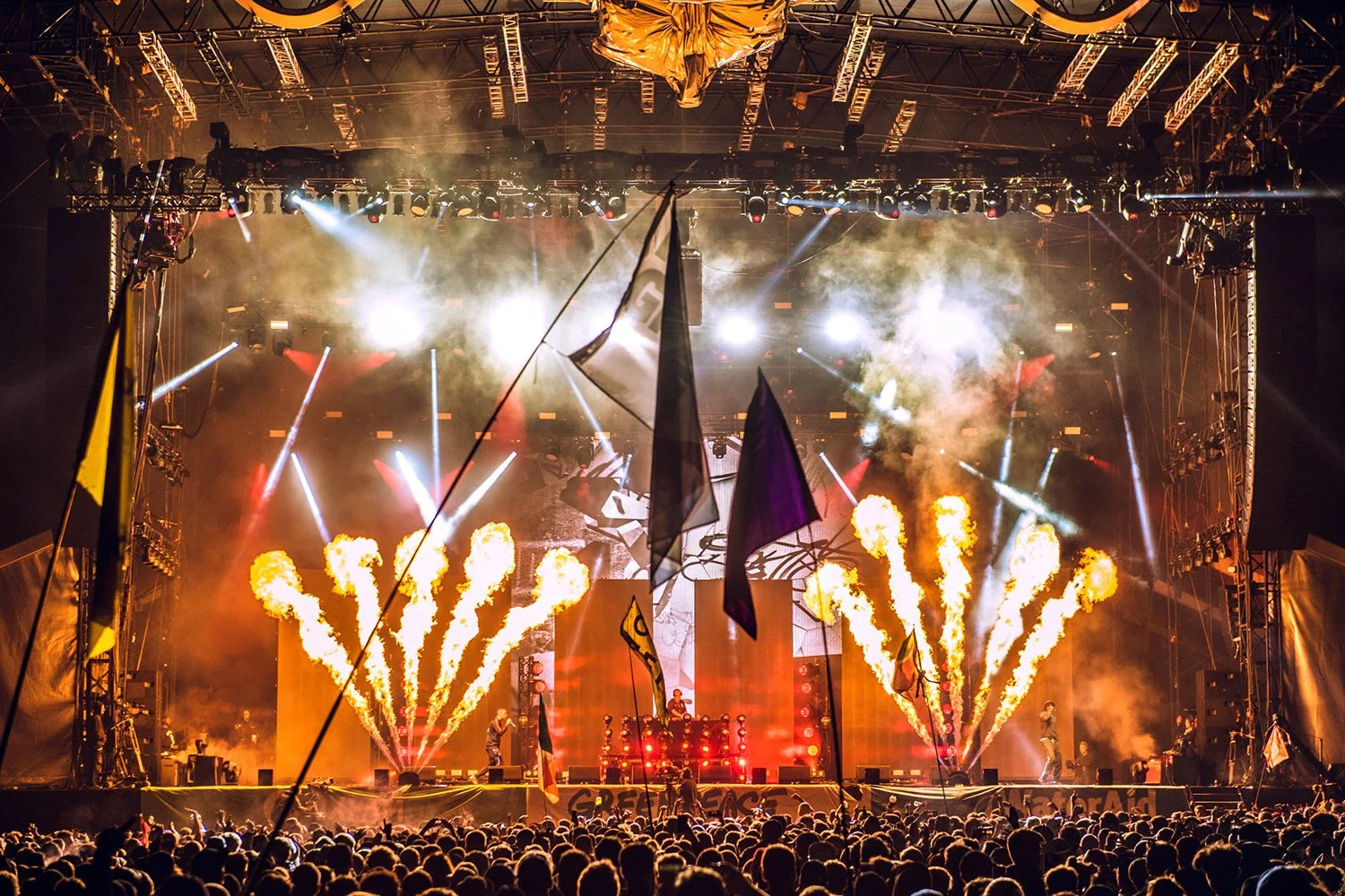 A concert stage with bright lights, pyrotechnics, and smoke, featuring a DJ or performer. The audience is visible at the bottom, waving flags and enjoying the show.