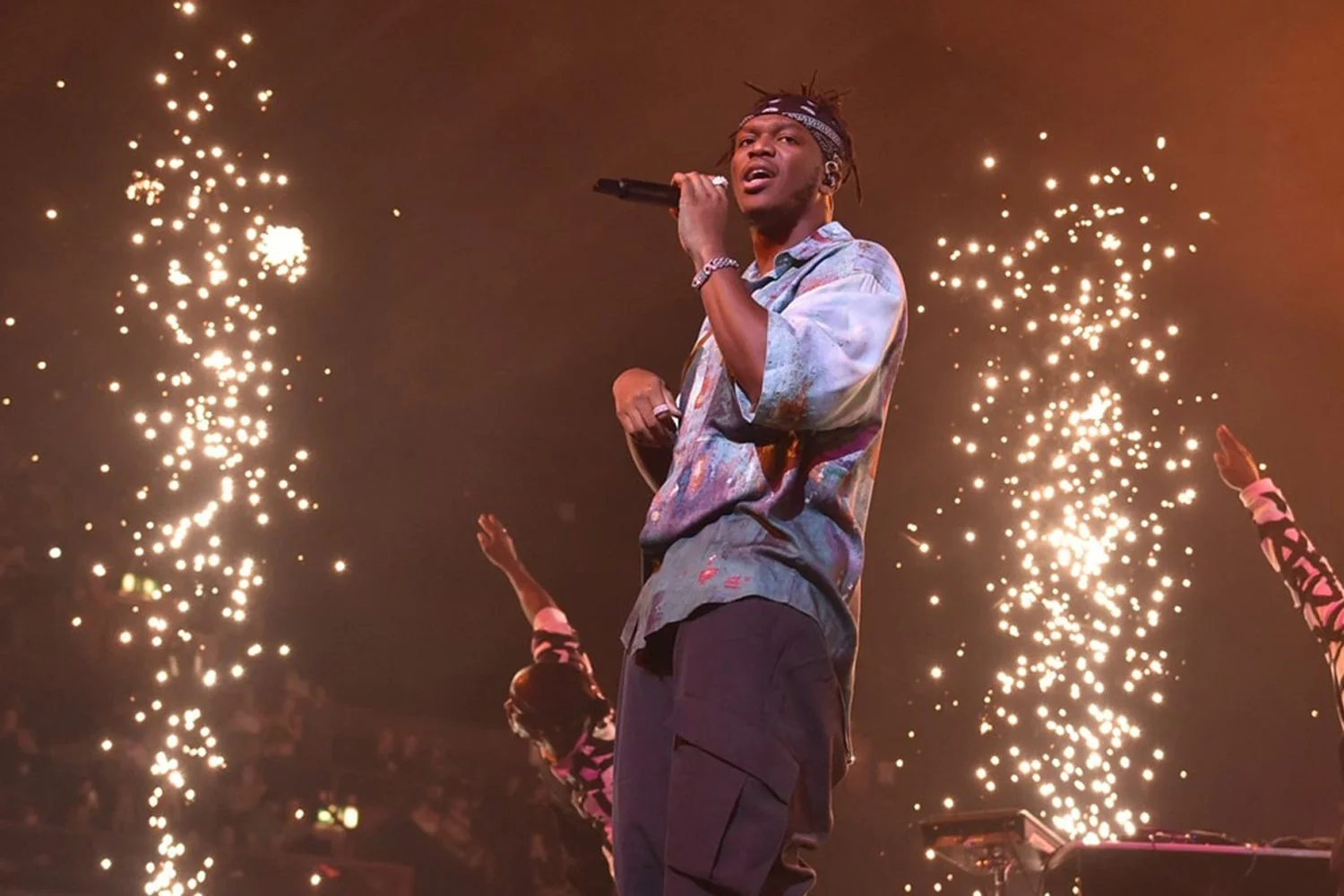 Performer on stage holding a microphone with fireworks or sparkles in the background during a concert or event.