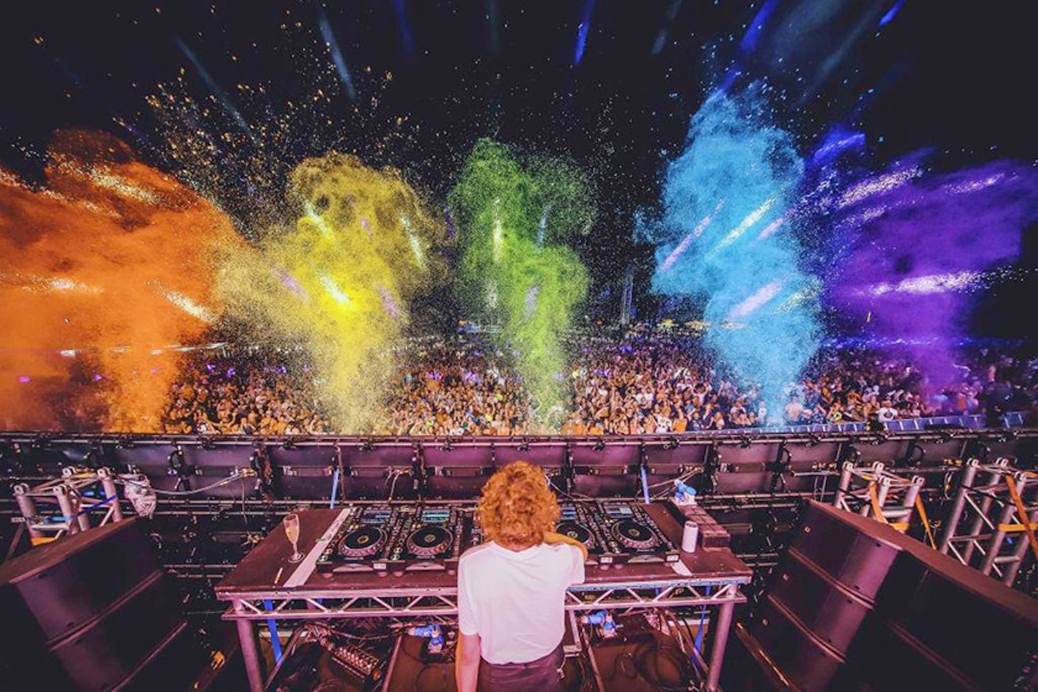 A DJ with curly hair standing behind a DJ setup on a stage at an outdoor party, with colorful fireworks illuminating the night sky and a large crowd of people dancing below.