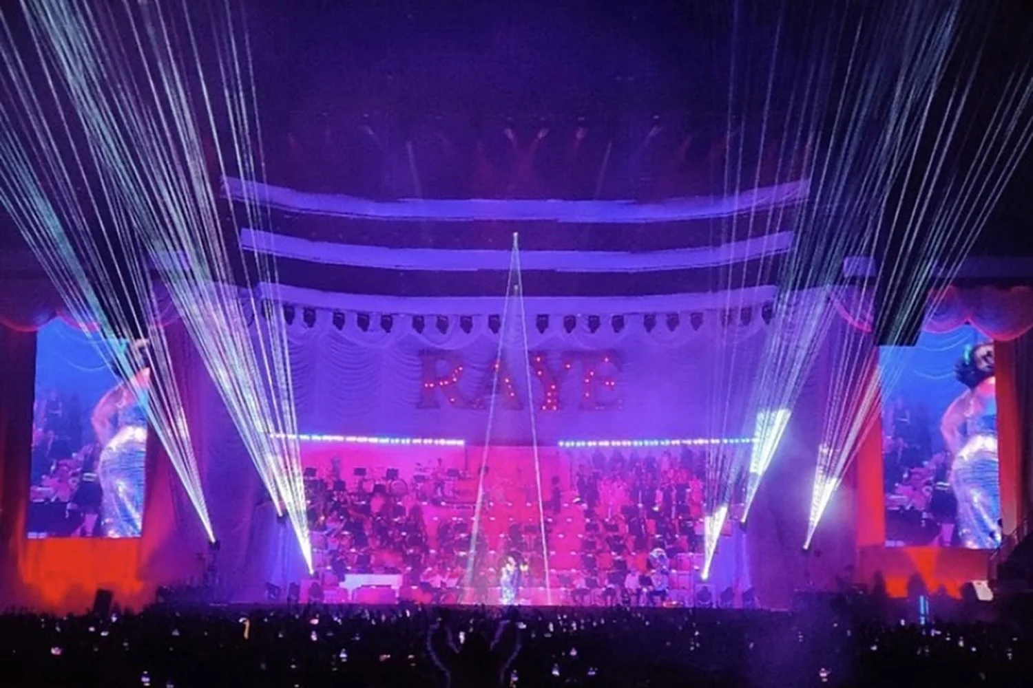 A stage with purple and pink lighting, laser beams, and dancers performing during a concert or show.