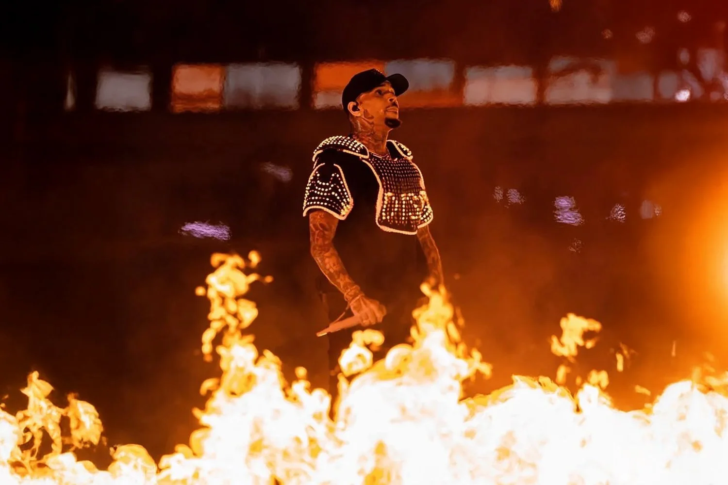 A man with tattoos standing amidst flames at night, wearing a black cap and clothing decorated with reflective studs.
