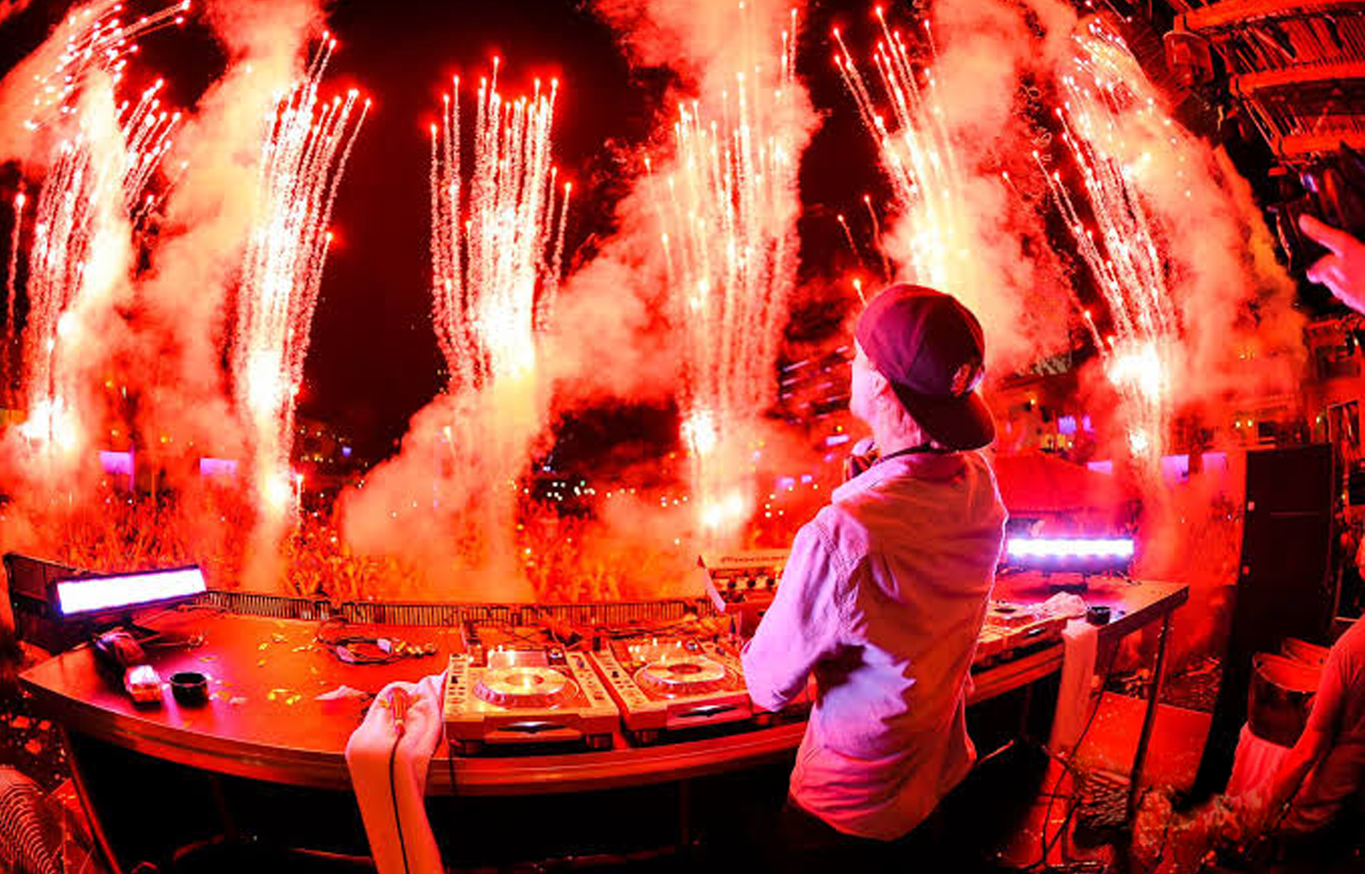 A DJ performing at a fireworks celebration at night, with bright red fireworks in the sky and a crowd in the background.