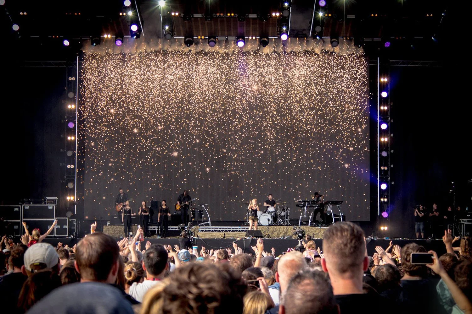 Music concert with a band performing on stage, large crowd watching, backdrop display of golden confetti or fireworks, stage lit with purple and white lights.