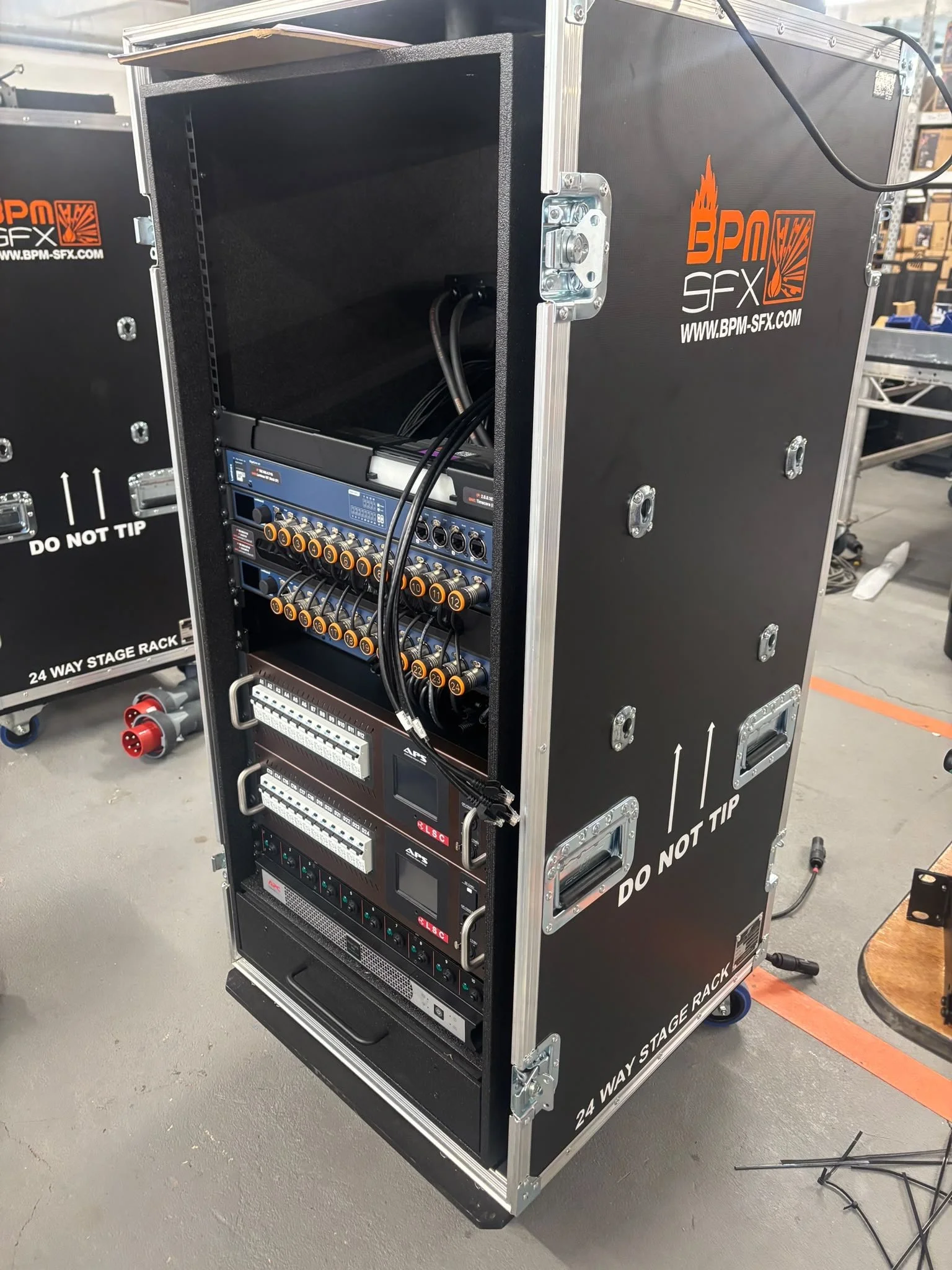 A black road case labeled '24 WAY STAGE RACK' contains audio equipment and cables, situated on a gray concrete floor in a warehouse or staging area.