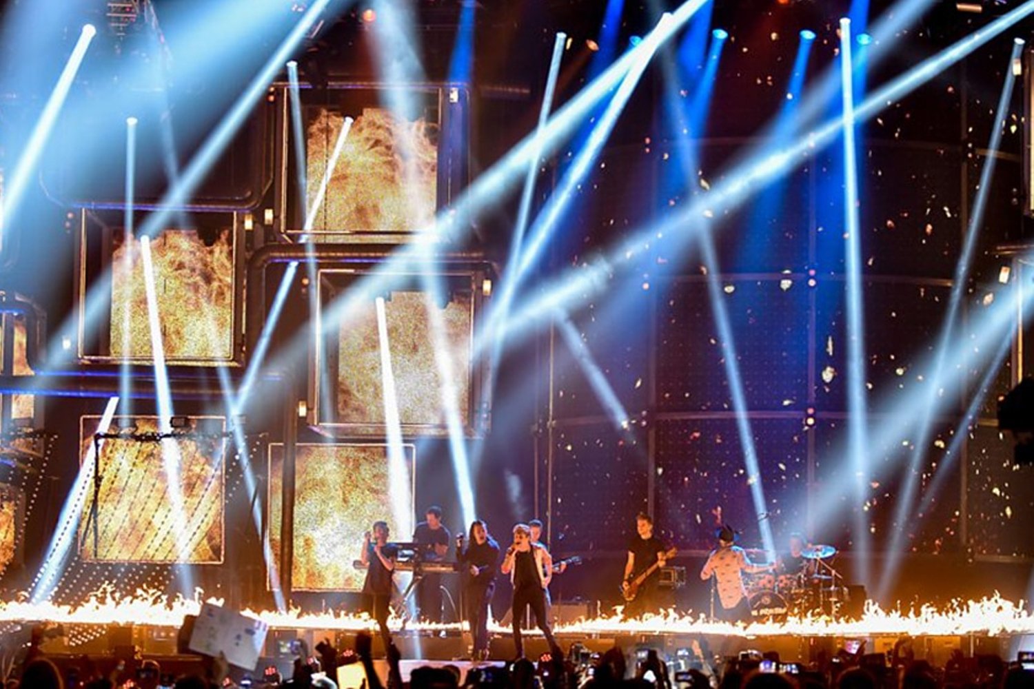 A live concert performance with a band on stage, illuminated by bright white and blue stage lights, with fiery effects and decorative screens in the background.