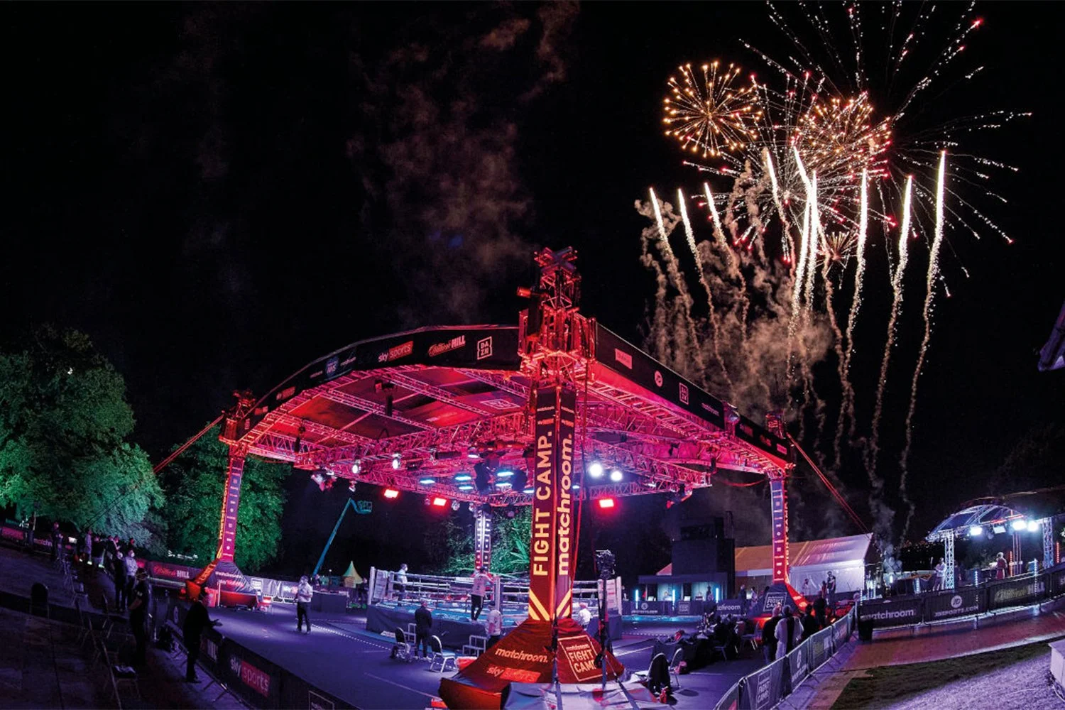 Nighttime outdoor boxing ring with red lighting and fireworks above, surrounded by spectators and seating area with event branding.