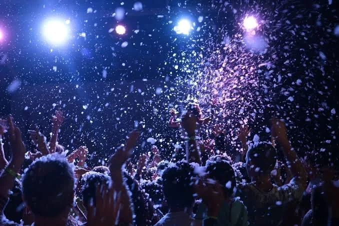 Crowd of people at a concert or event with bright lights and foam or confetti being thrown into the air.